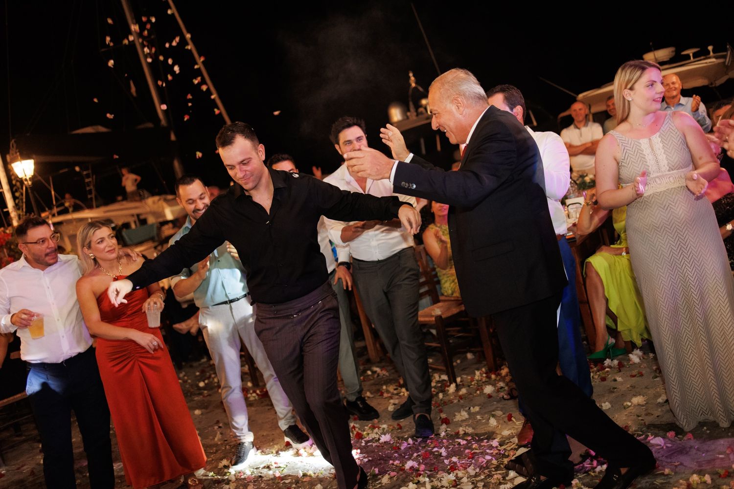 Wedding guests performing traditional Greek line dance outdoors at a nighttime reception.