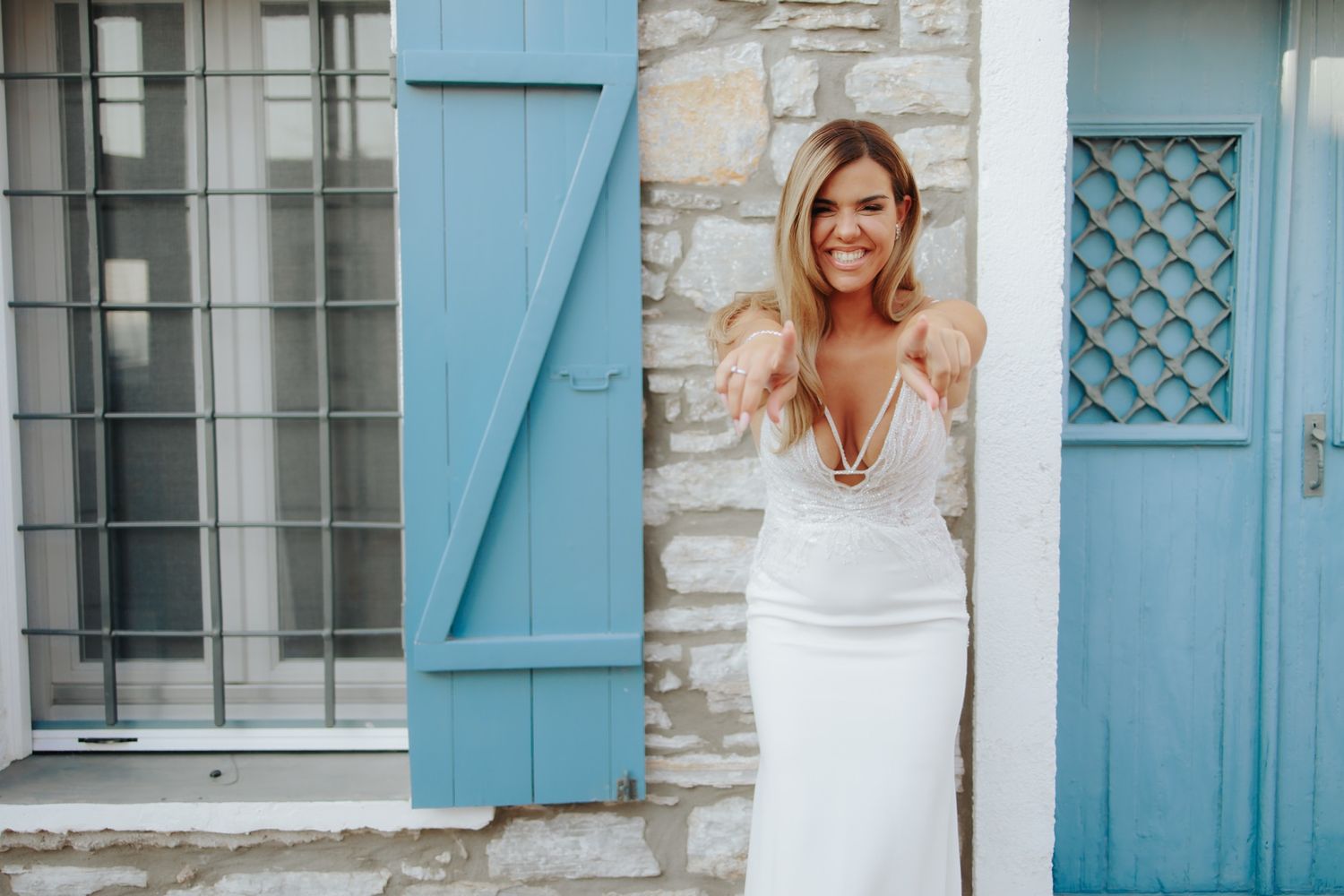 Smiling bride in white wedding dress pointing at camera beside blue door on stone building.