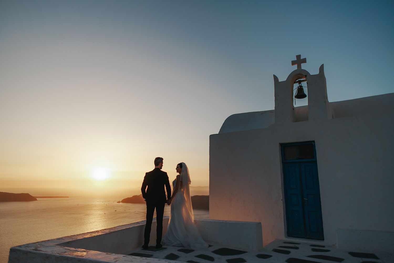 Silhouettes stand by a white Greek chapel with a bell tower overlooking the Mediterranean Sea at sunset.