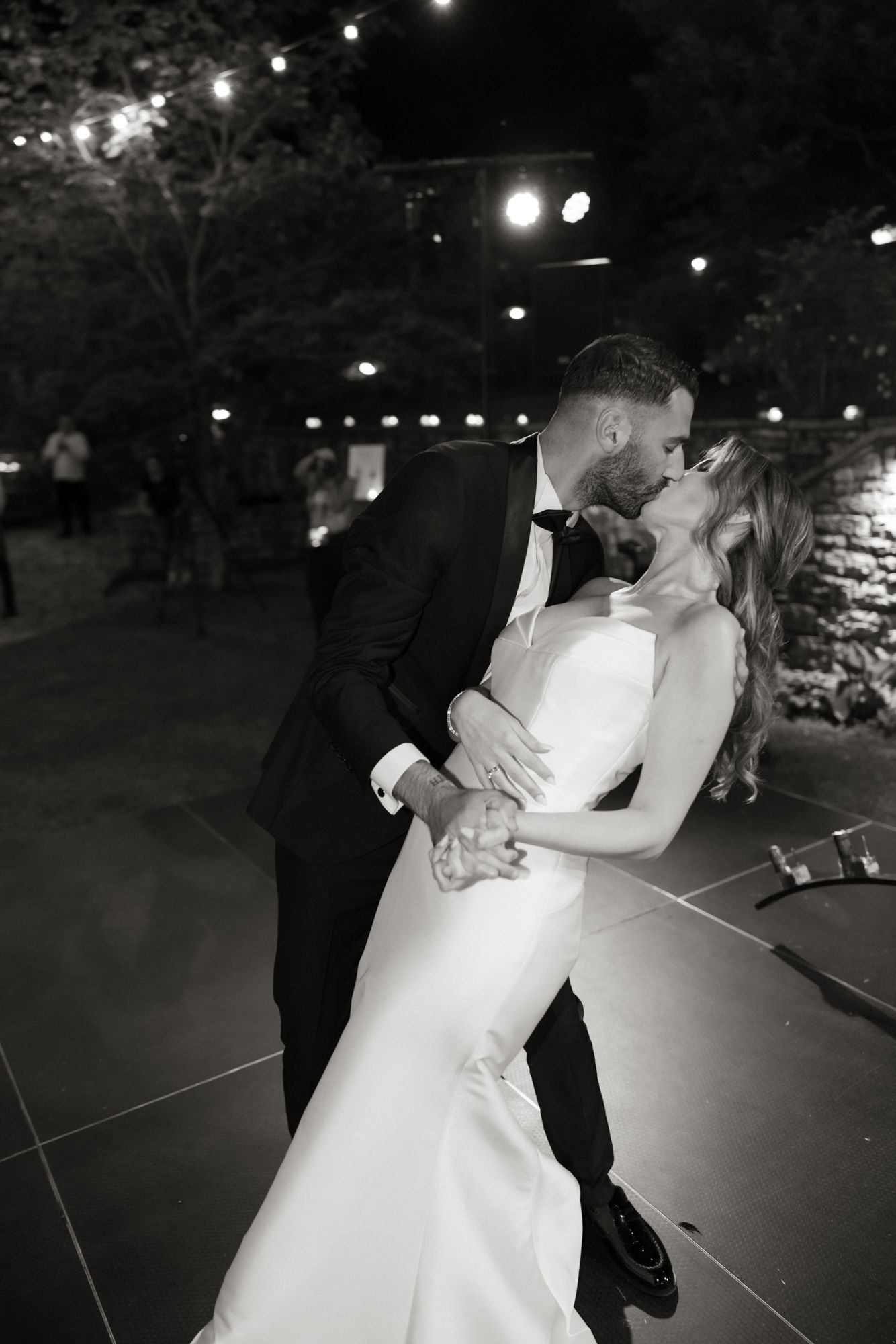 Bride and groom share passionate kiss on dance floor at night in black and white wedding photo.