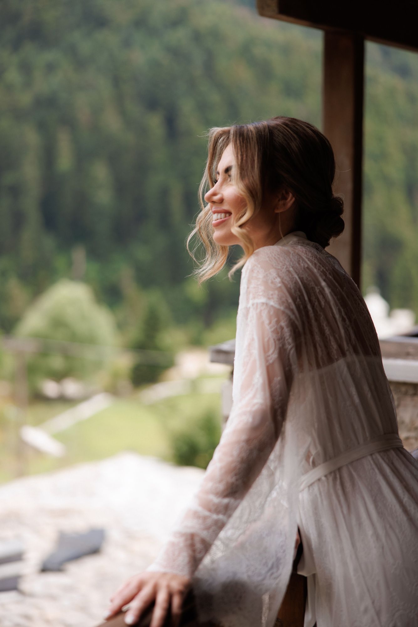 Joyful bride in white lace robe smiling on balcony with scenic green mountain forest backdrop.
