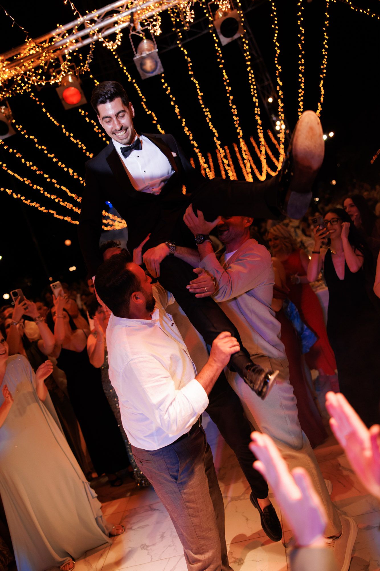 Groom lifted by groomsmen on wedding dance floor surrounded by fairy lights and cheering guests at night.