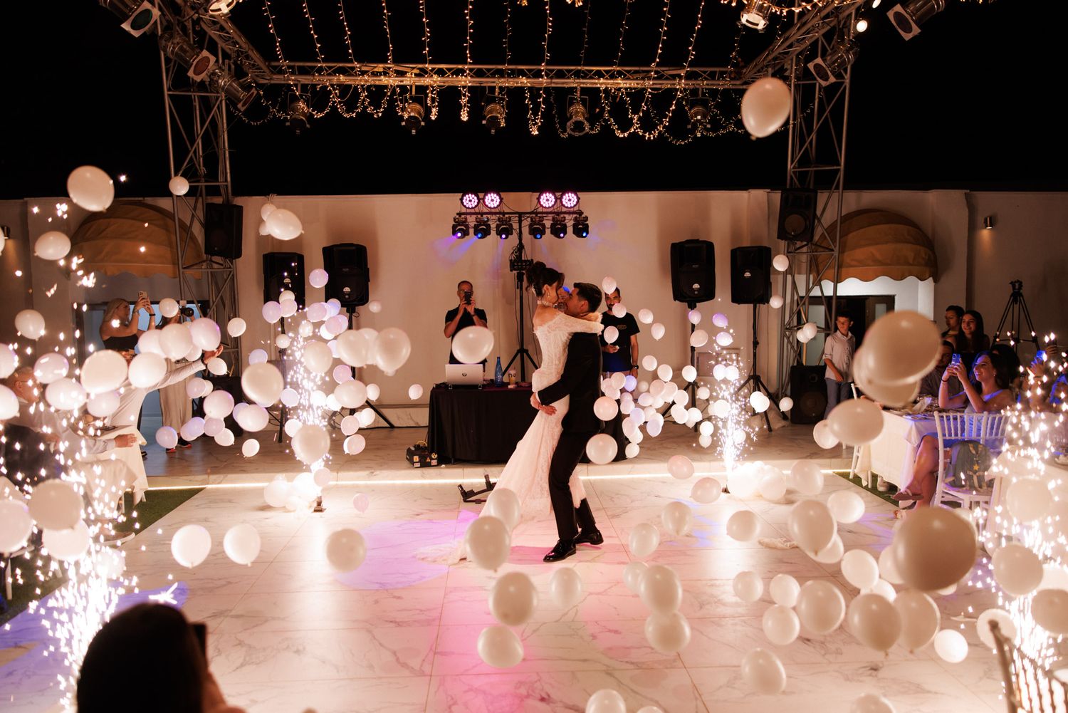Bride and groom share first dance surrounded by white balloons and sparkling lights on an elegant white dance floor.