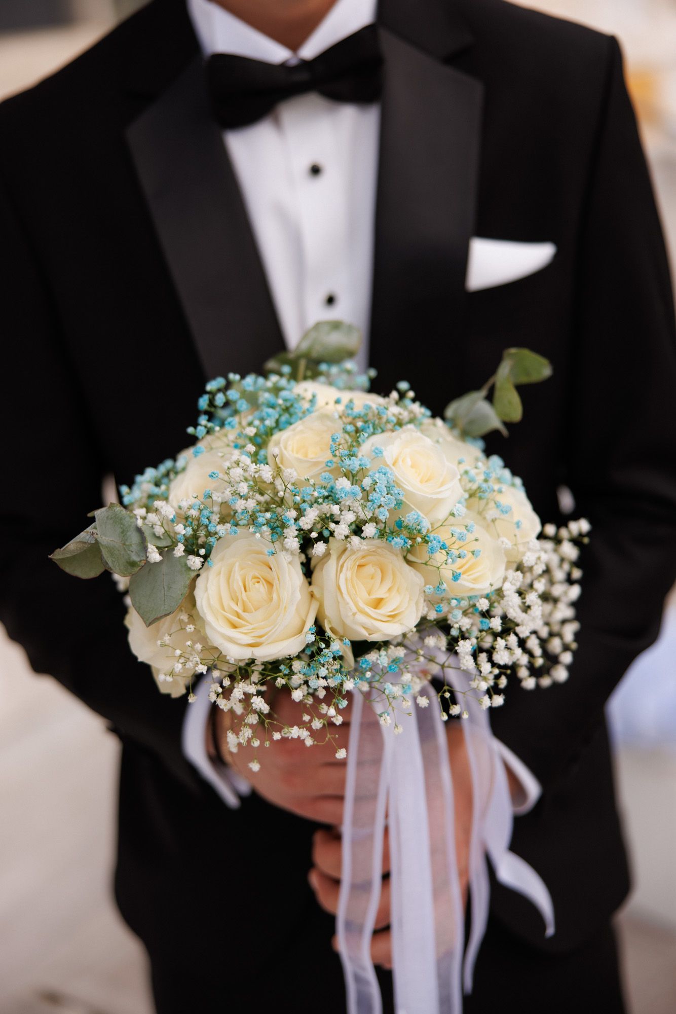 Groom in black tuxedo holding a bridal bouquet of white roses and blue baby's breath with white ribbons.