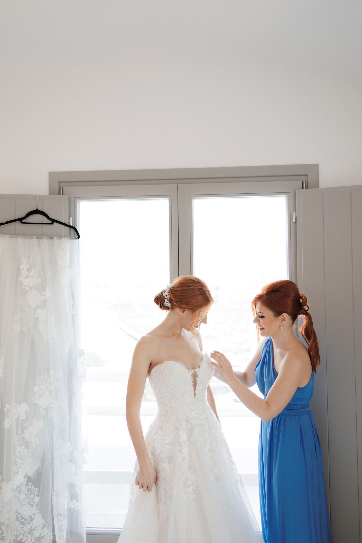 Bridesmaid in blue dress helping bride in strapless lace gown prepare near a bright window.