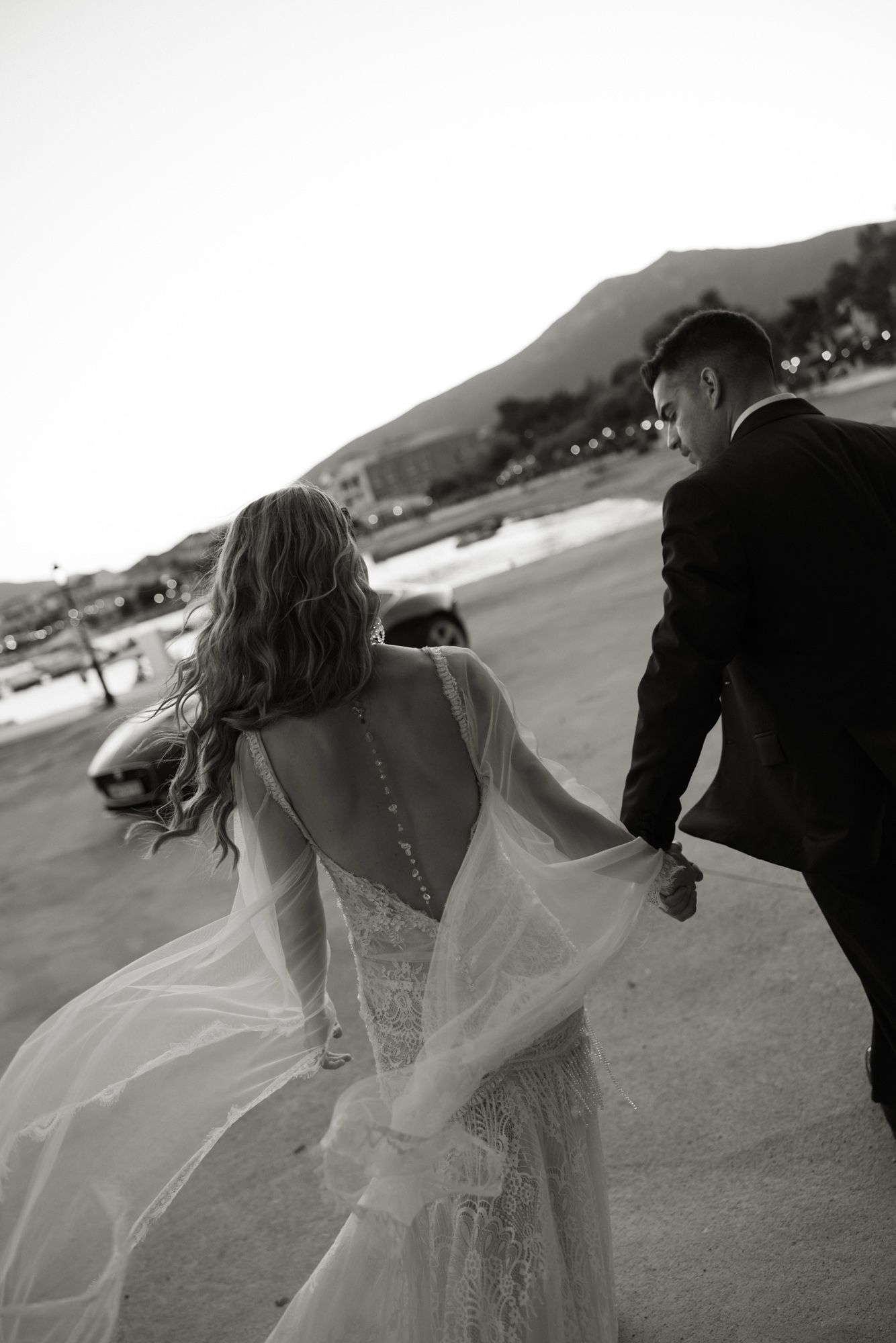 Black and white photo of bride and groom holding hands walking near waterfront at dusk.