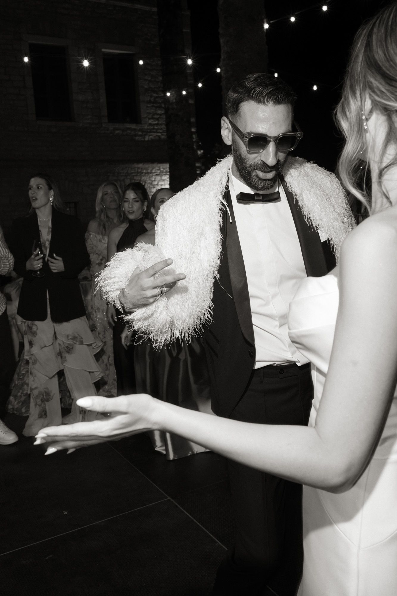 Groom in tuxedo with white feather stole and sunglasses dancing at wedding reception in black and white photo.
