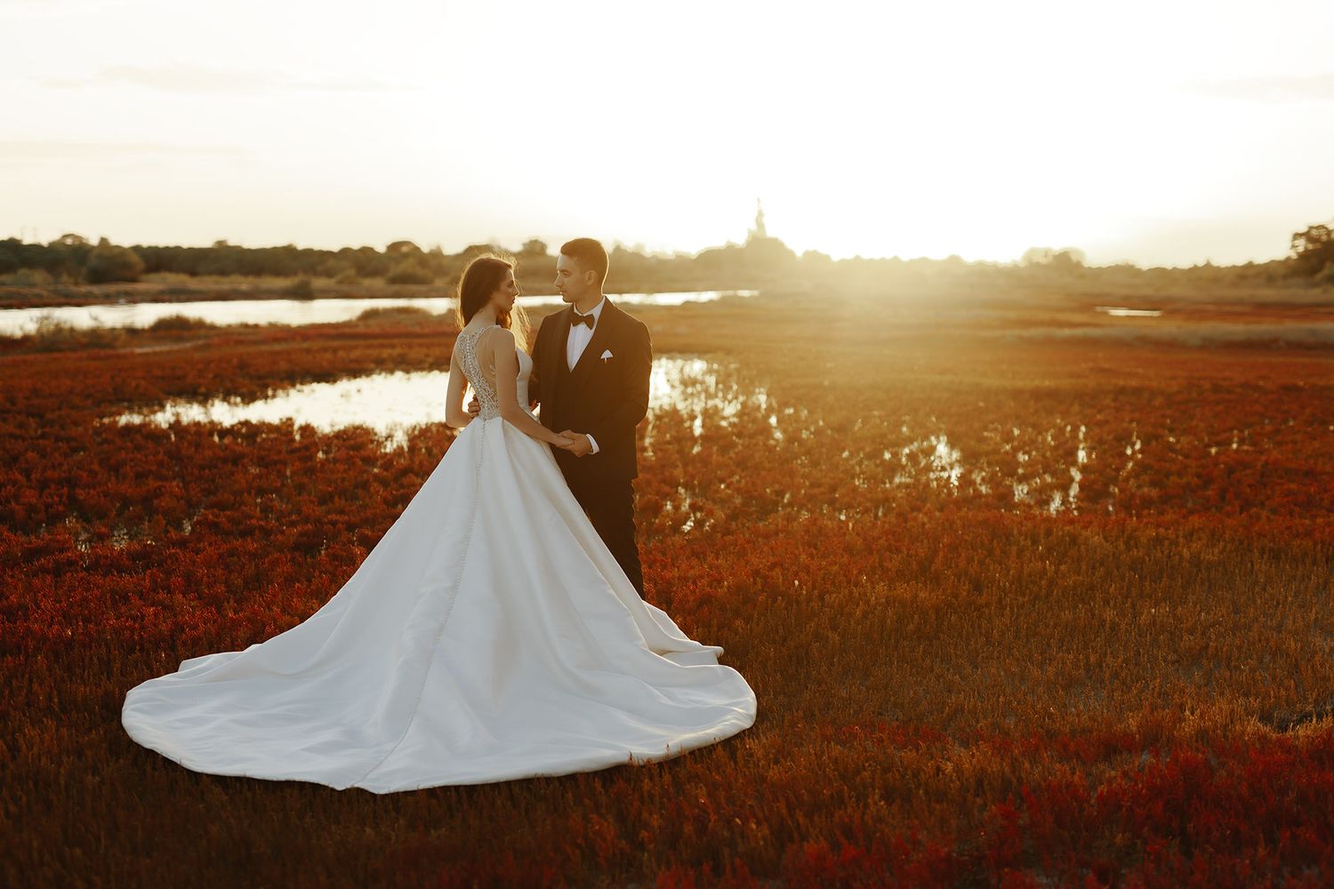 A couple in wedding attire embraces at sunset near a marsh, with the bride's white gown trailing elegantly behind her.