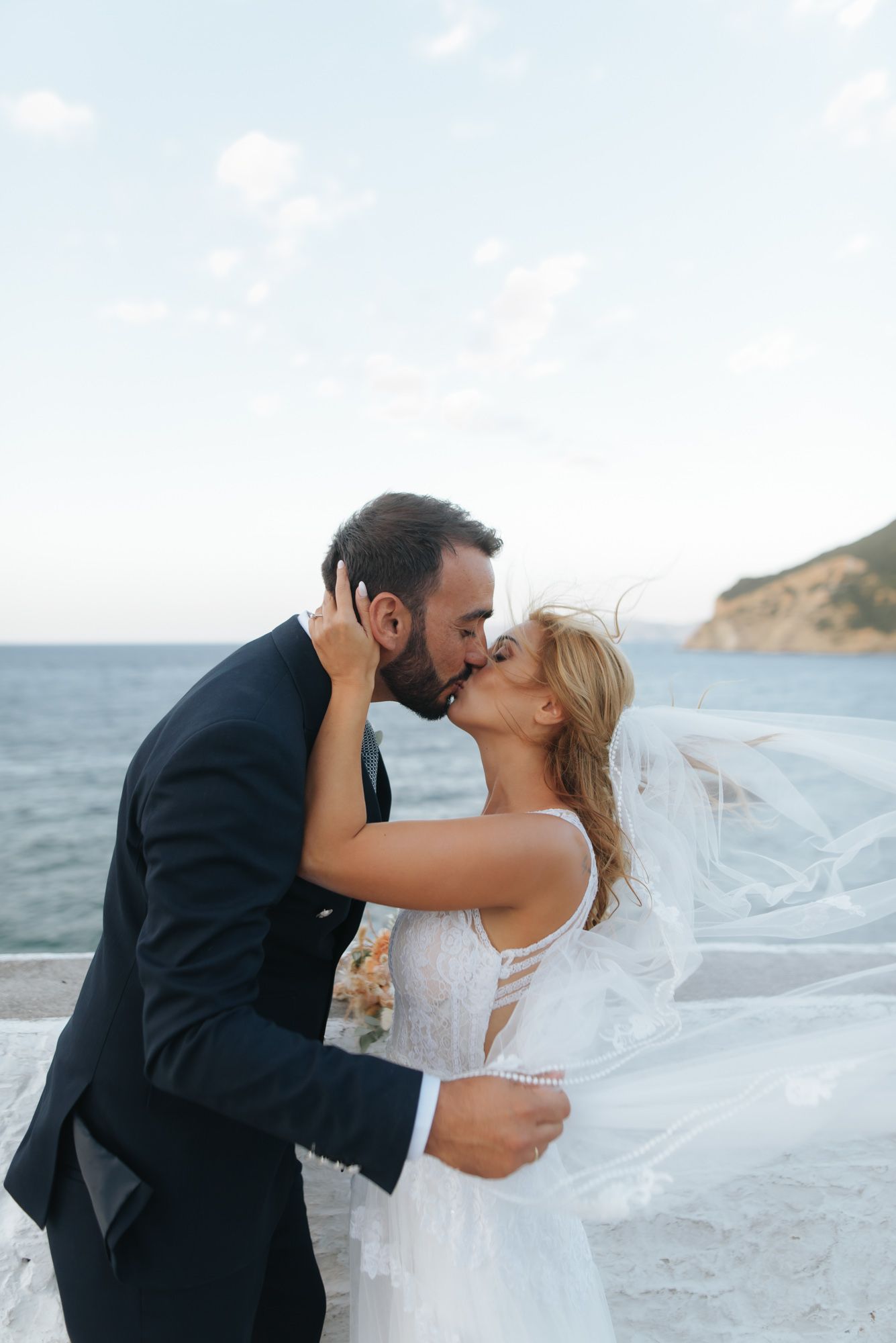 Bride and groom share a passionate kiss with flowing veil and ocean backdrop on their wedding day.