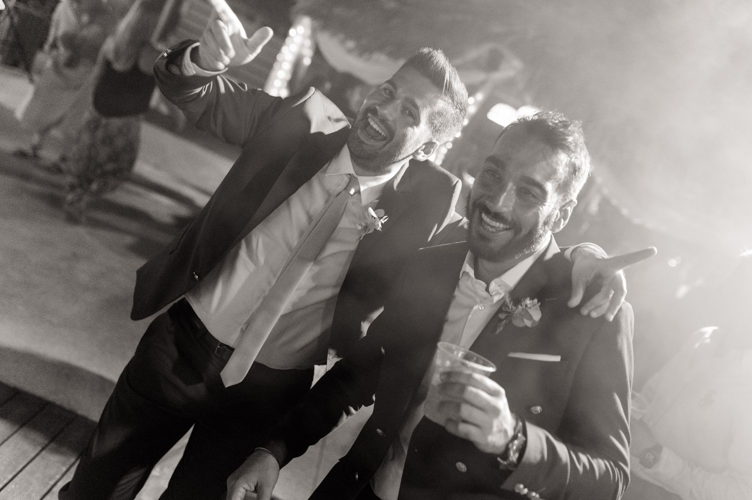Two joyful grooms in suits celebrate together at their wedding reception in a black and white photo.