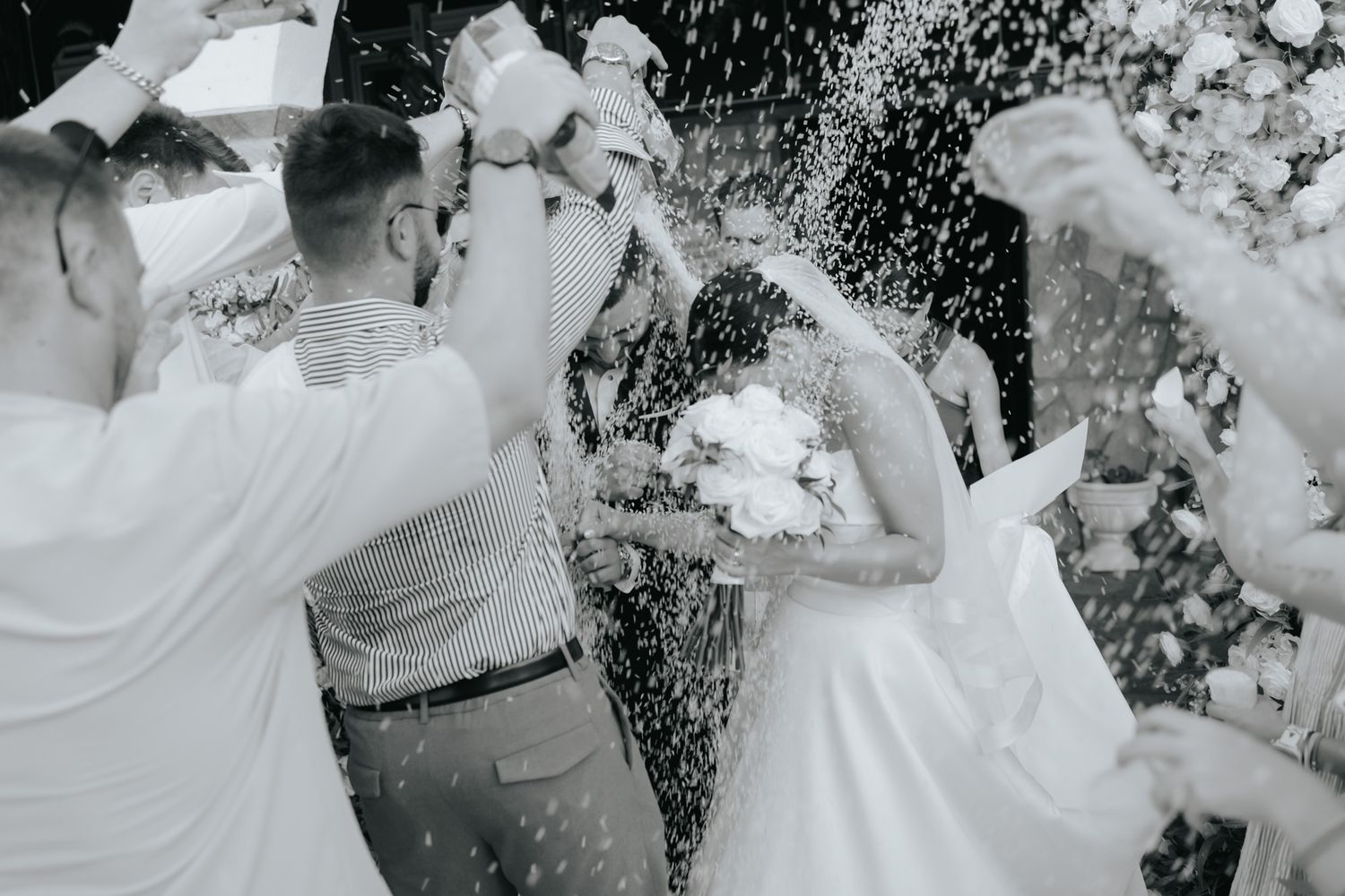 Black and white photo of newlyweds being showered with rice by guests during a joyful wedding celebration.