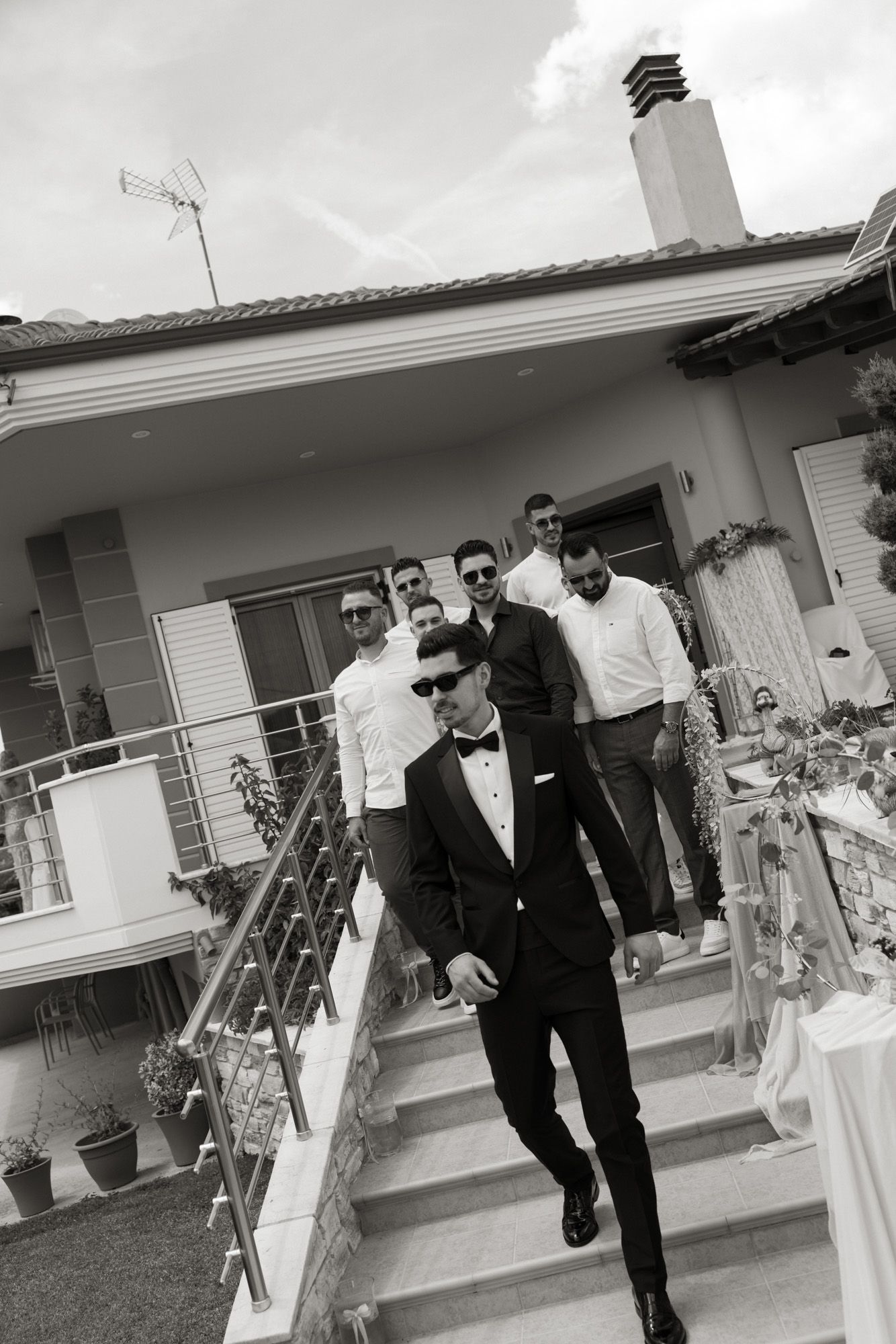 Groom in tuxedo leads groomsmen down outdoor stairs at wedding venue, black and white photo.