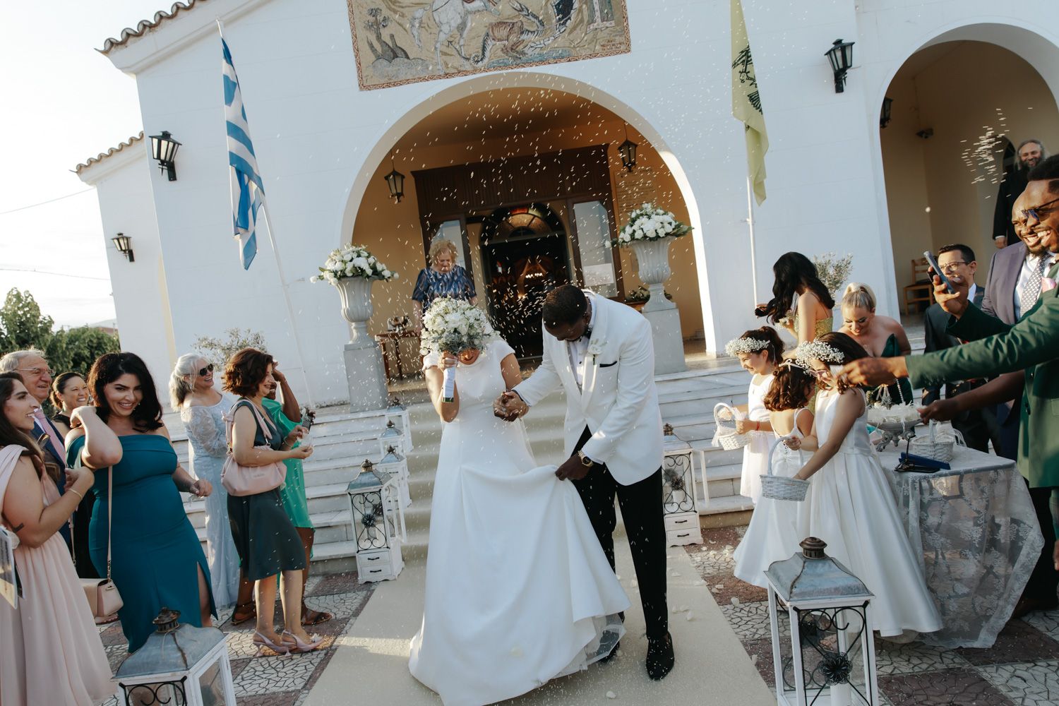 Bride and groom exit Greek Orthodox church ceremony surrounded by guests throwing confetti outdoors.