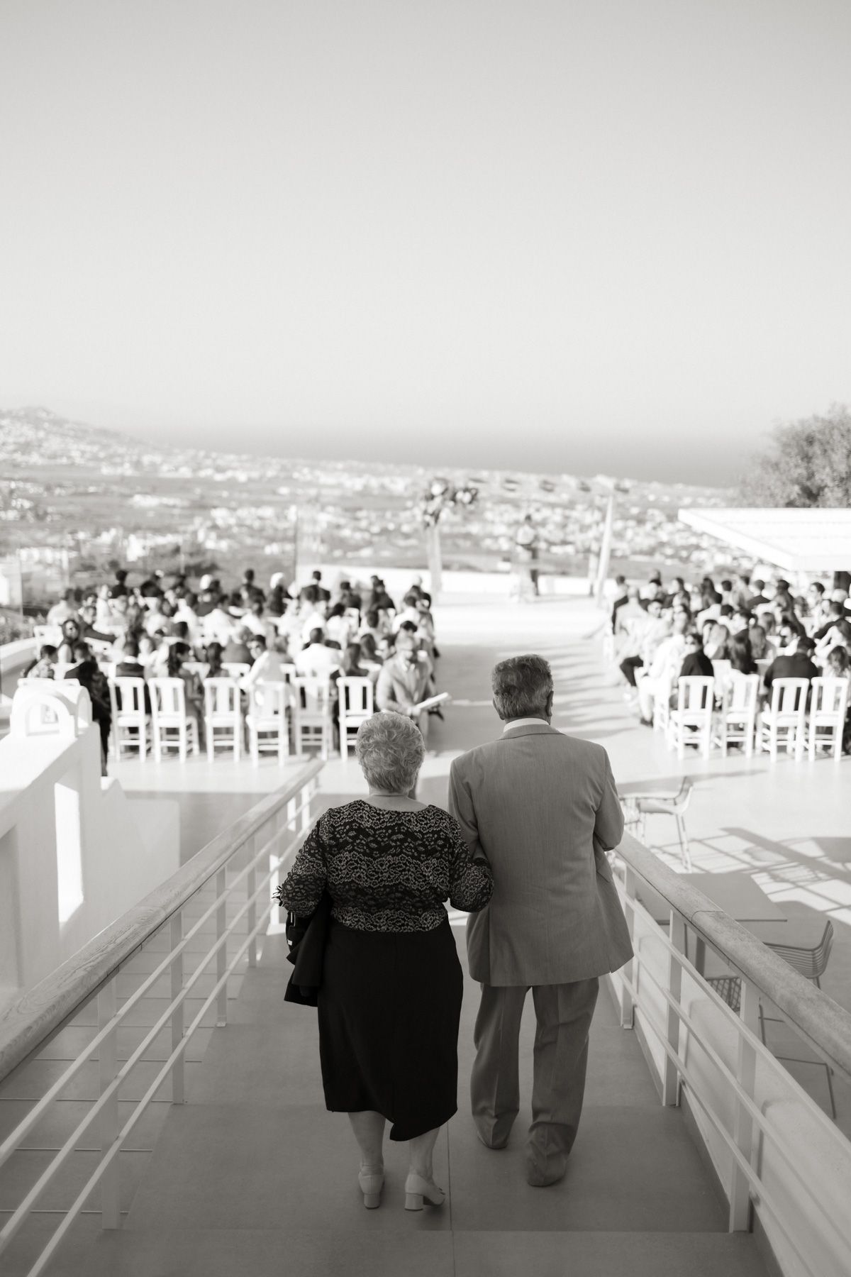 Black and white photo of elderly couple walking toward outdoor wedding ceremony with sea view.