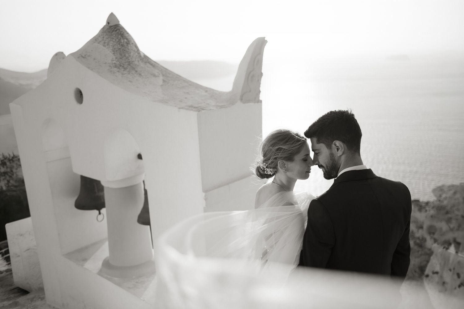 Black and white photo of bride and groom sharing intimate moment near a white chapel overlooking the sea.