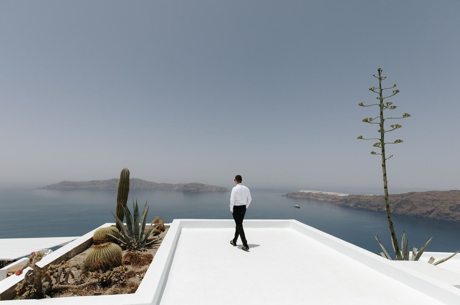 A person stands on a white rooftop terrace overlooking the blue waters and cliffs of Santorini, Greece.