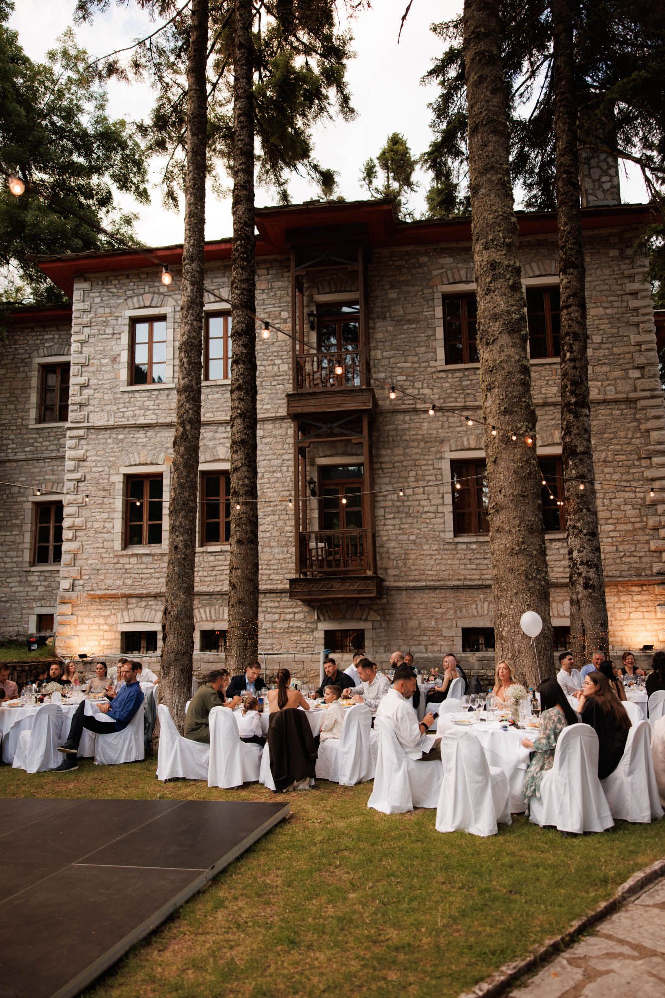Outdoor wedding reception with guests at white-draped tables beside a stone manor surrounded by tall pine trees.