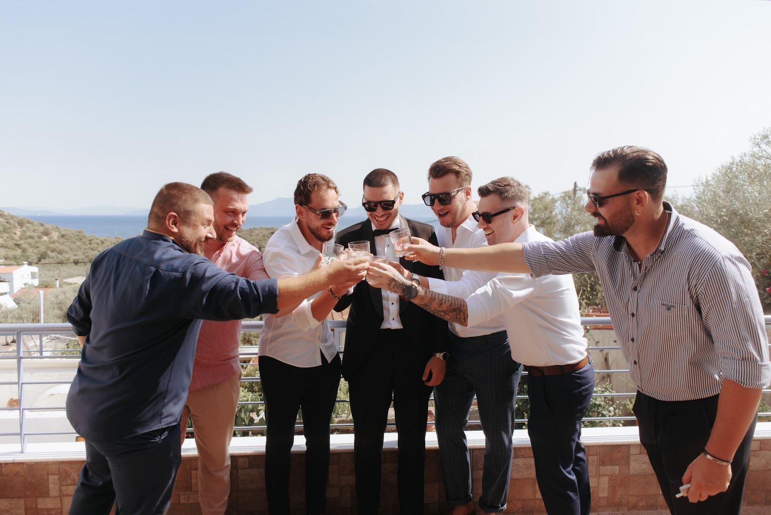 Group of groomsmen toasting drinks together on a sunny outdoor terrace with scenic views.