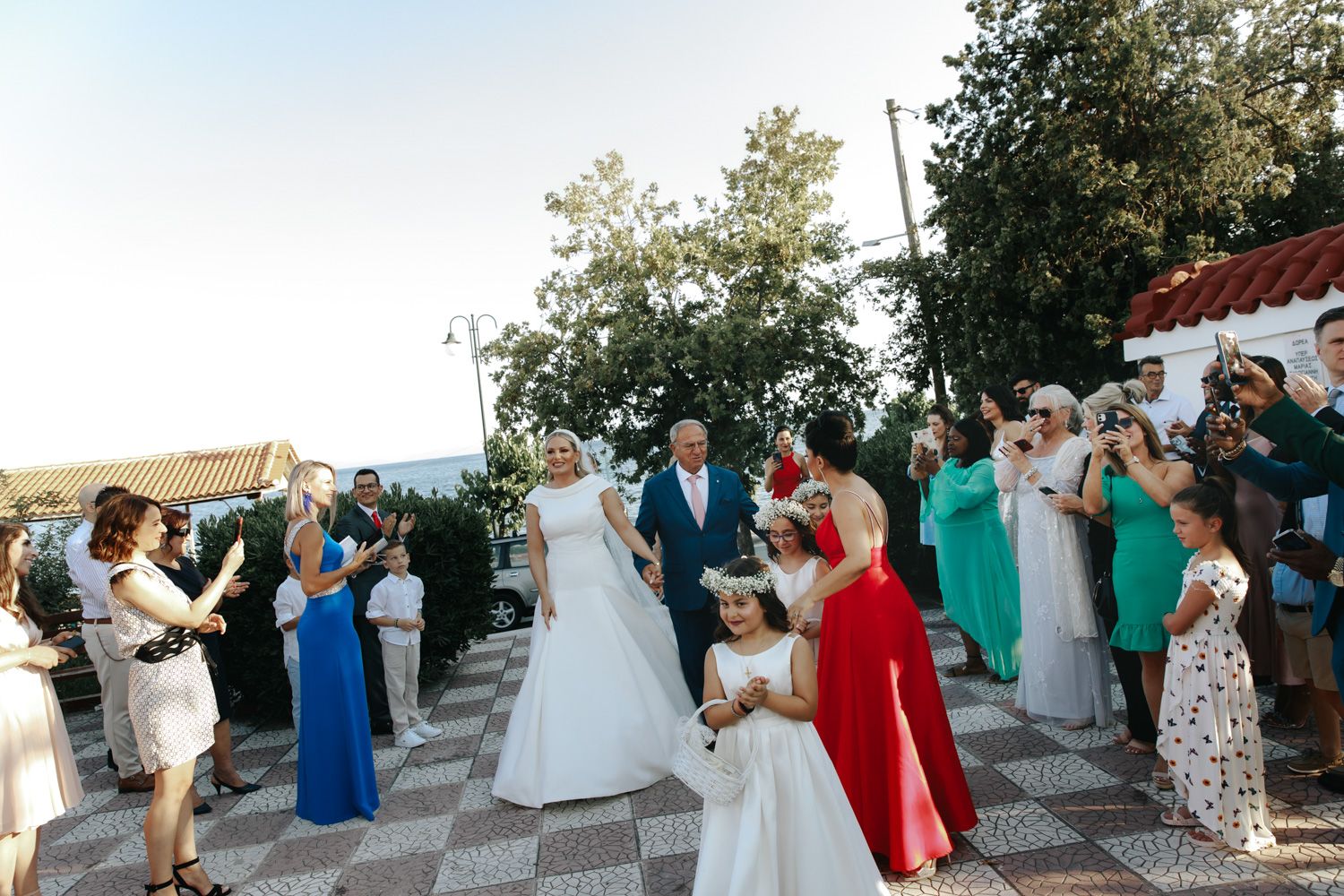Bride in white gown walks with flower girl at outdoor wedding ceremony while guests celebrate and applaud.