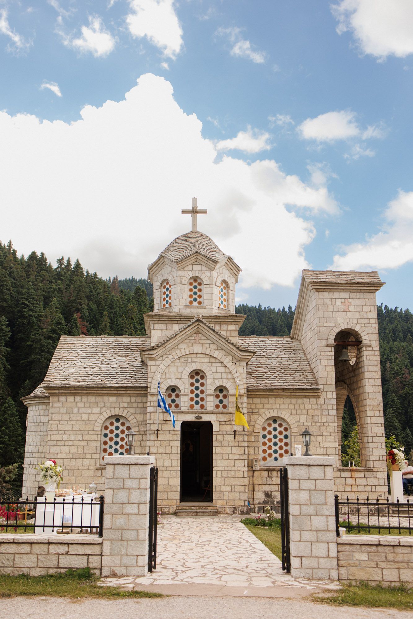 Small Orthodox stone church with a cross and flags surrounded by pine trees under blue sky.