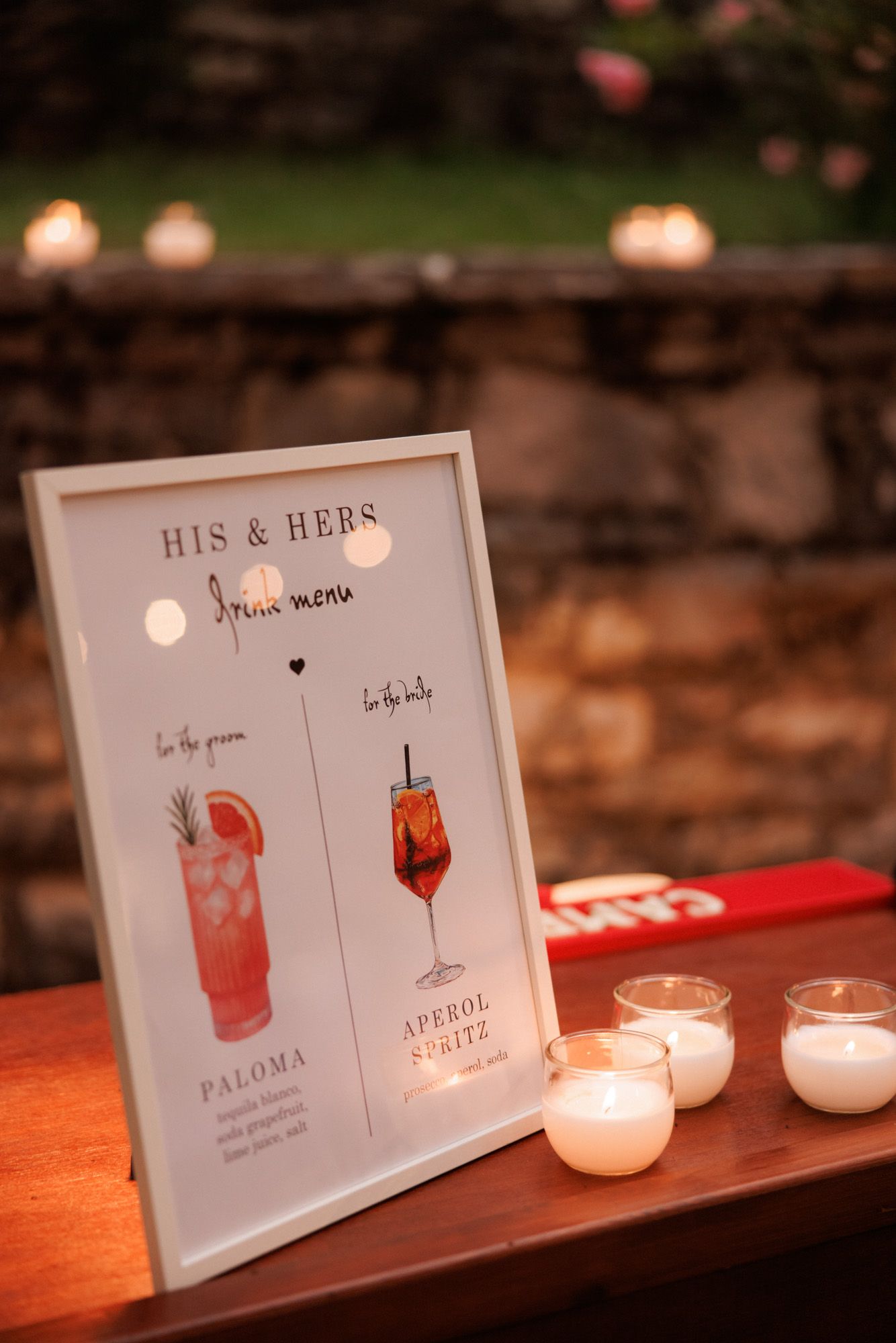 His and Hers drink menu sign featuring Paloma and Aperol Spritz cocktails displayed with candles at wedding.