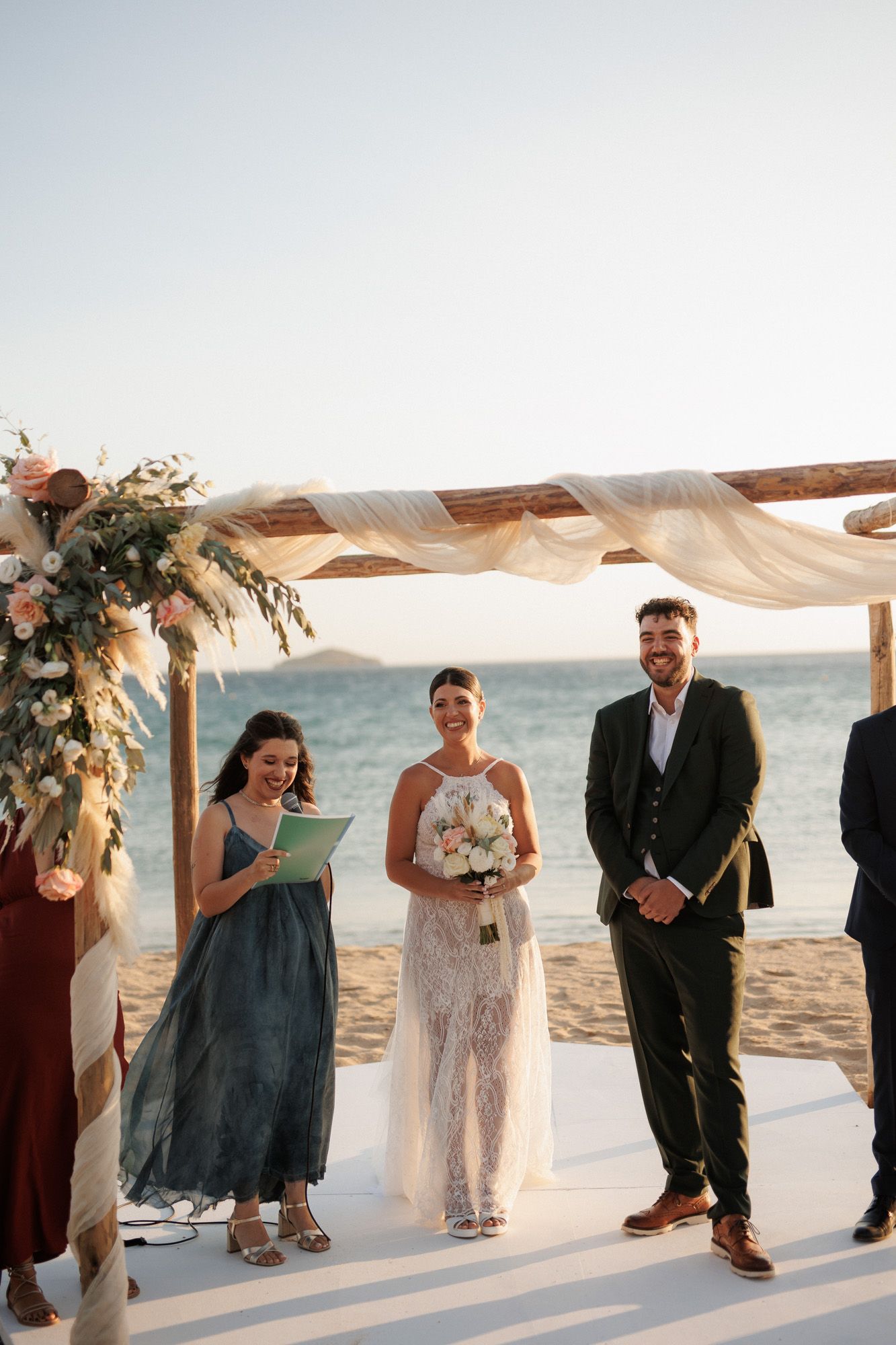Bride and groom smile during outdoor beach wedding ceremony under floral wooden arch by the sea.