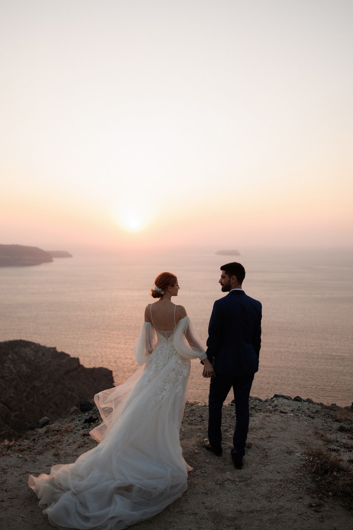 Wedding couple stands on Santorini cliff at sunset, bride in elegant white dress with long train, groom in navy suit.