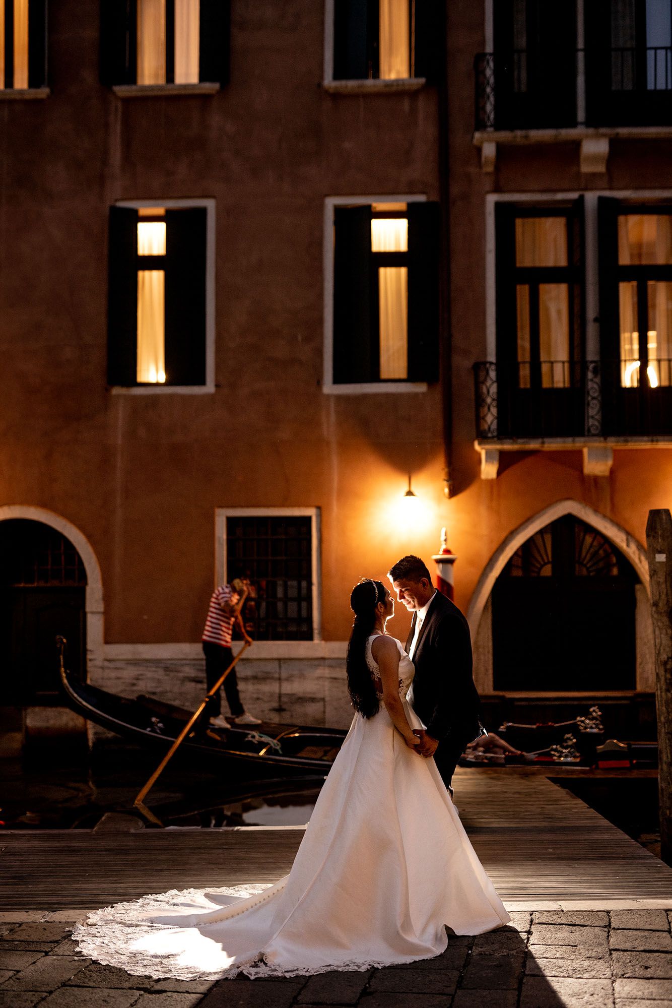 A romantic wedding couple stands on a Venice canal at night with illuminated buildings and gondolas in the background.