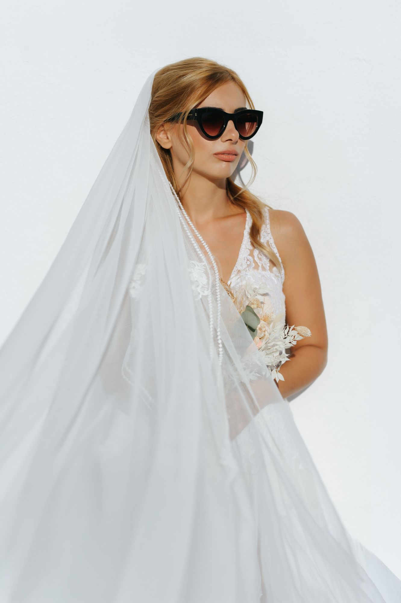 Stylish bride in white lace gown and veil wearing black cat-eye sunglasses holding a bridal bouquet.