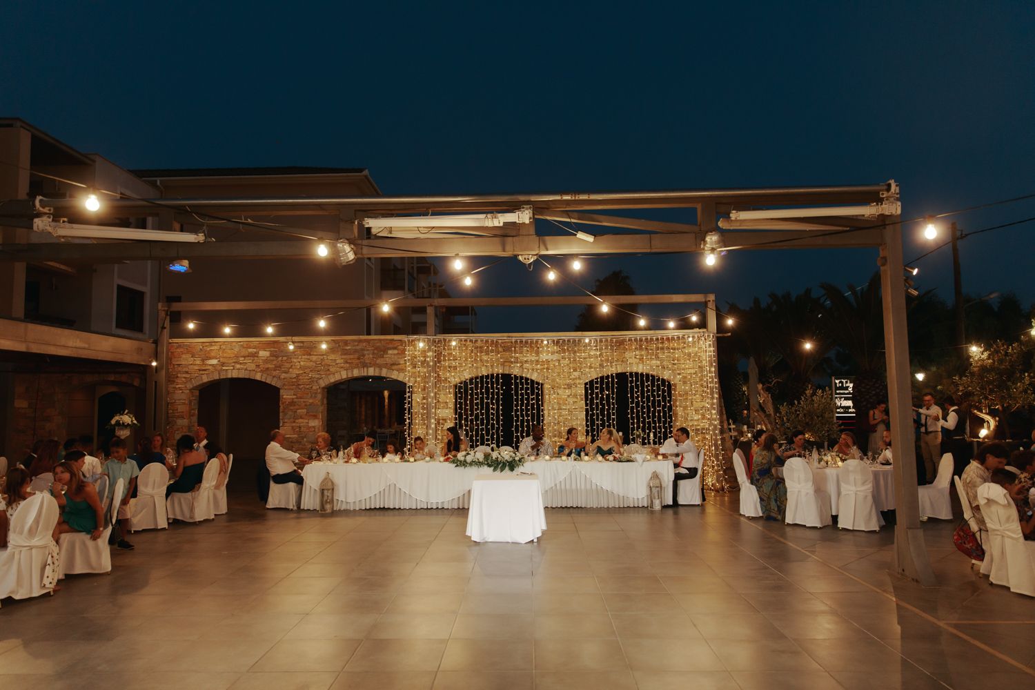 Outdoor wedding reception at night with string lights, brick walls, and guests seated at decorated tables.