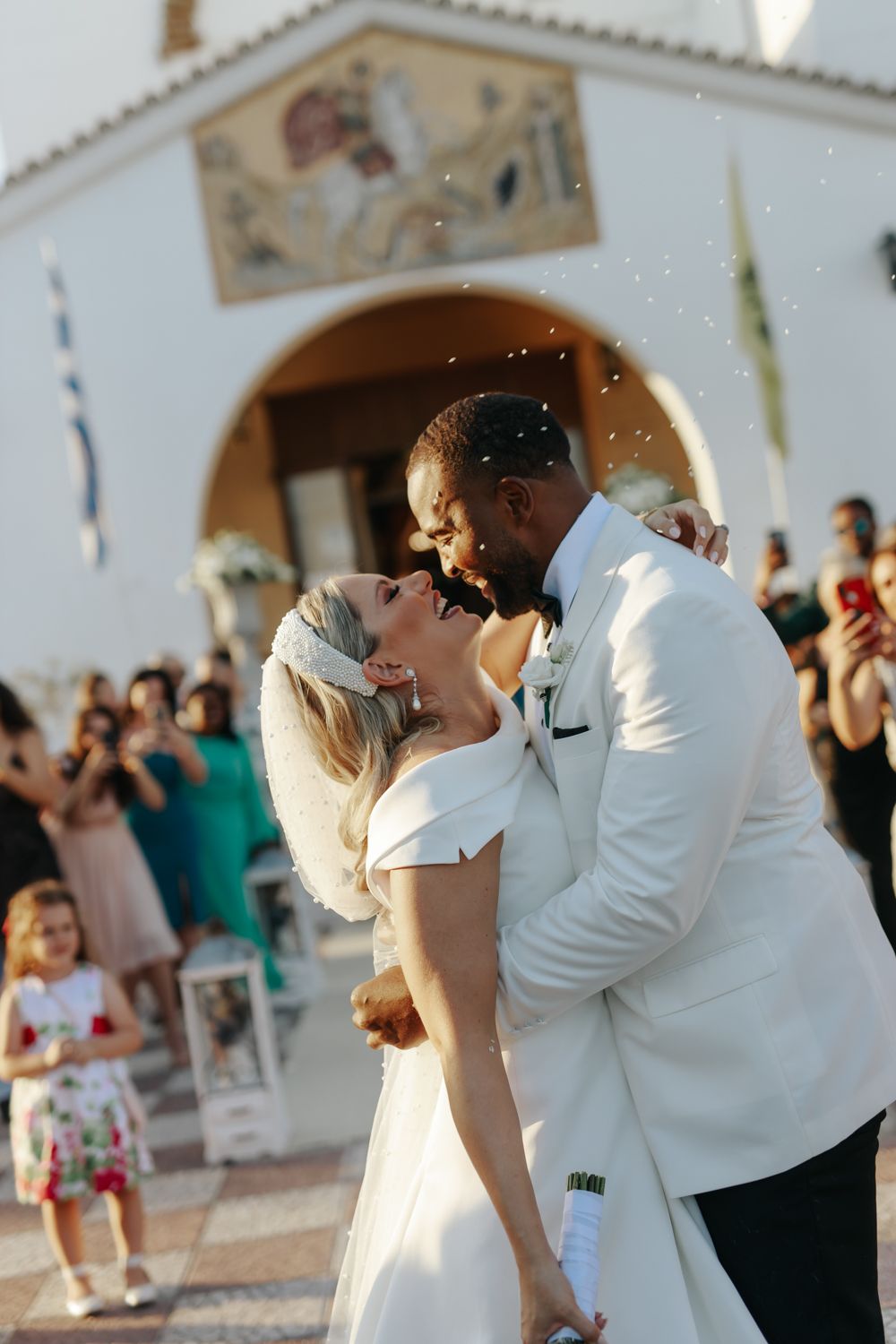 Bride and groom share first kiss outside white chapel with guests celebrating at outdoor wedding ceremony.