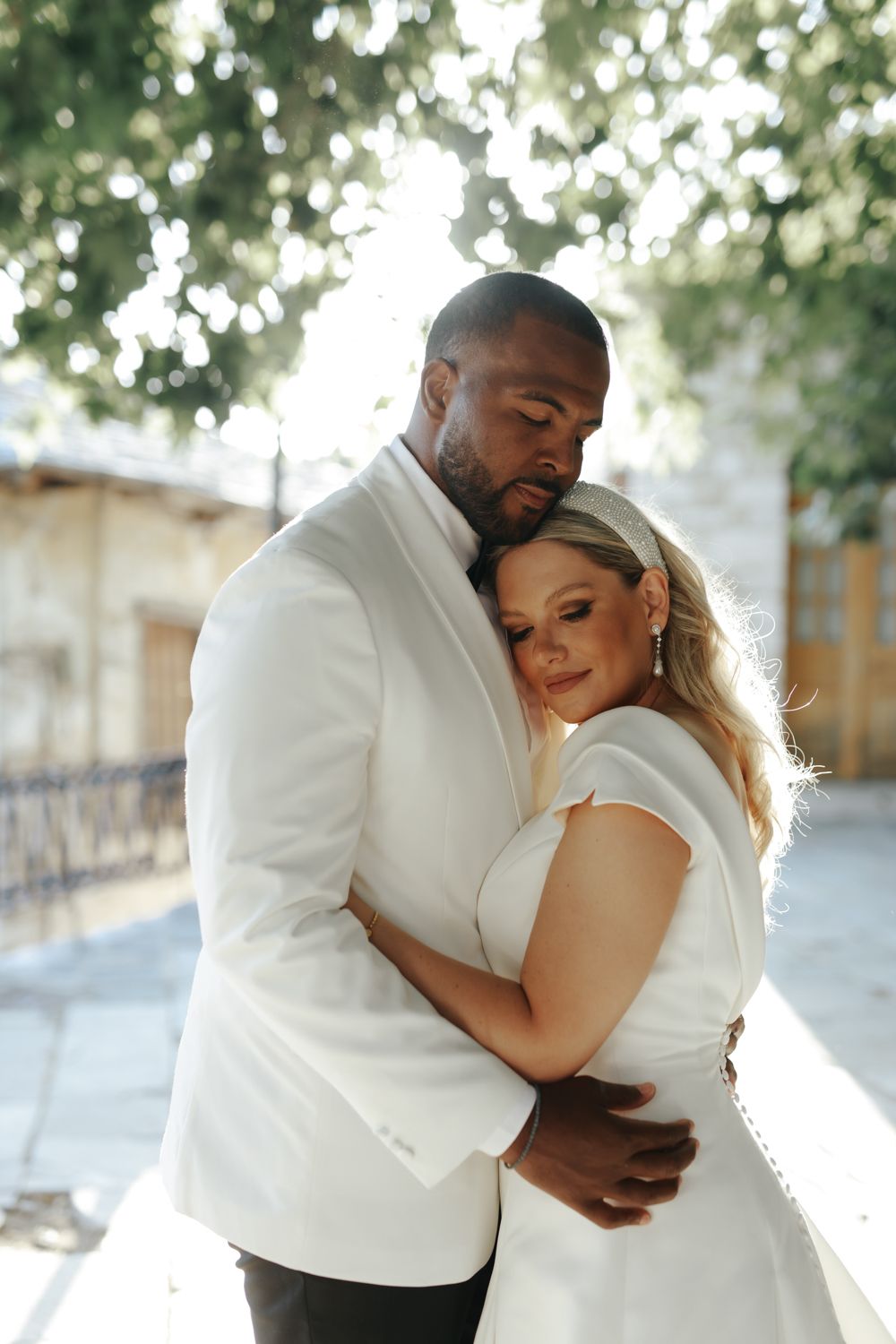 A couple in white wedding attire embraces tenderly outdoors, with the bride smiling softly against the groom's chest.