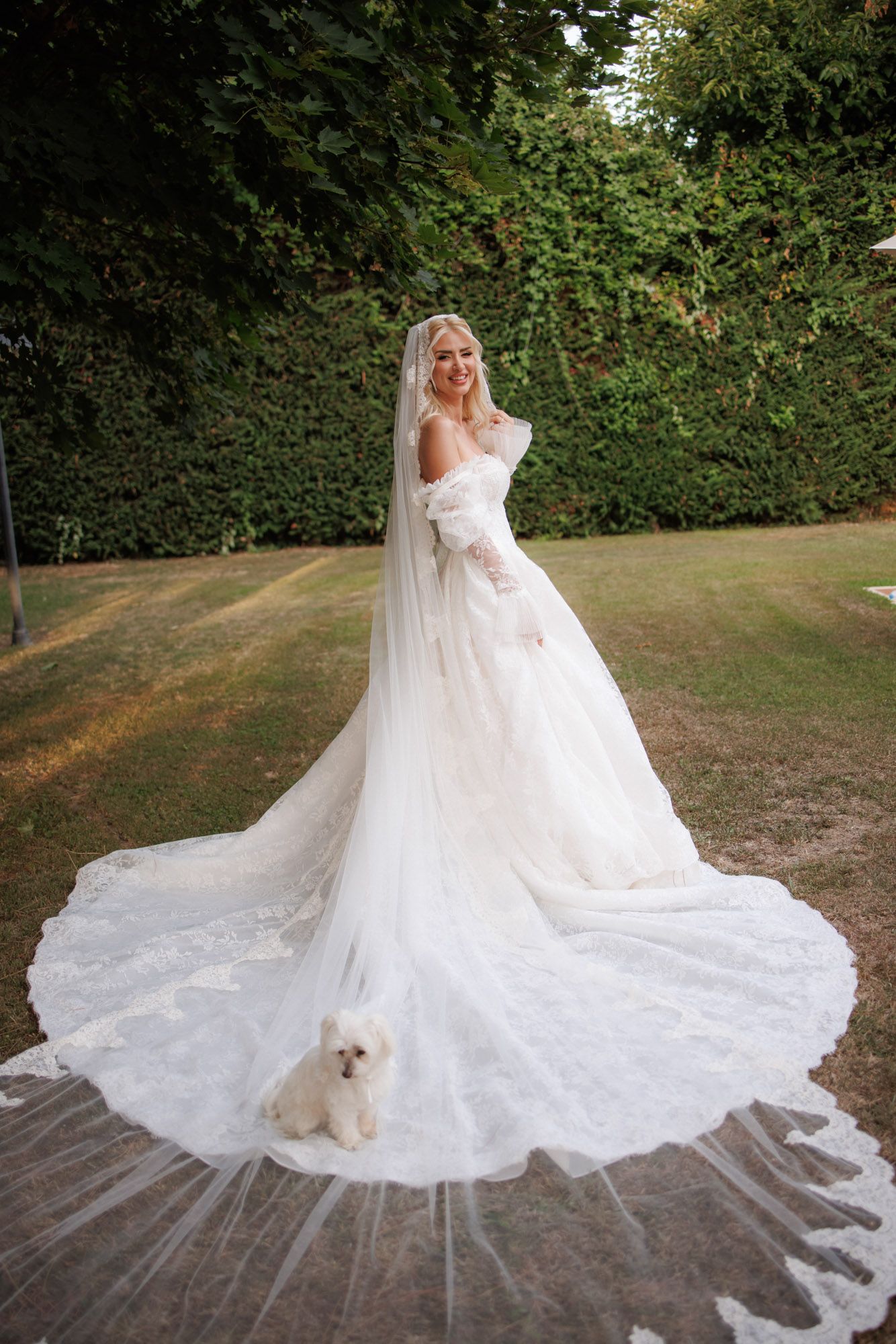 Radiant bride in off-shoulder lace wedding gown with cathedral veil poses outdoors with a small white fluffy dog.