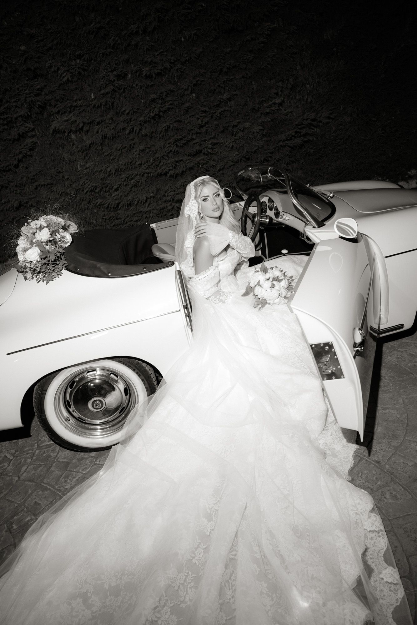 Black and white photo of blonde bride in voluminous gown posing beside classic vintage white convertible car.
