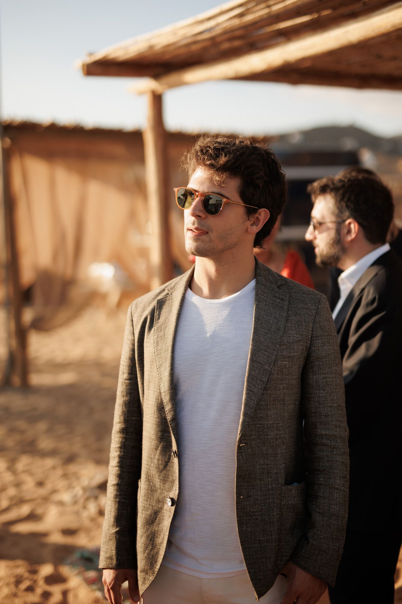 Stylish young man in gray blazer and sunglasses stands outdoors at a sunny beach event.