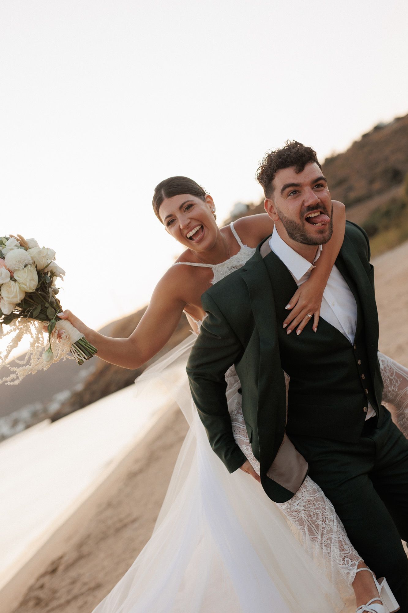 Laughing bride holds bouquet while groom gives her a piggyback ride on sunny beach.
