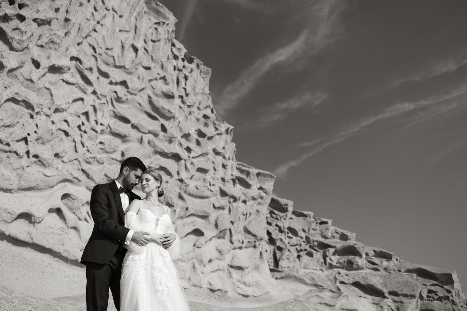 Black and white portrait of bride and groom embracing beside ancient textured stone wall under open sky.