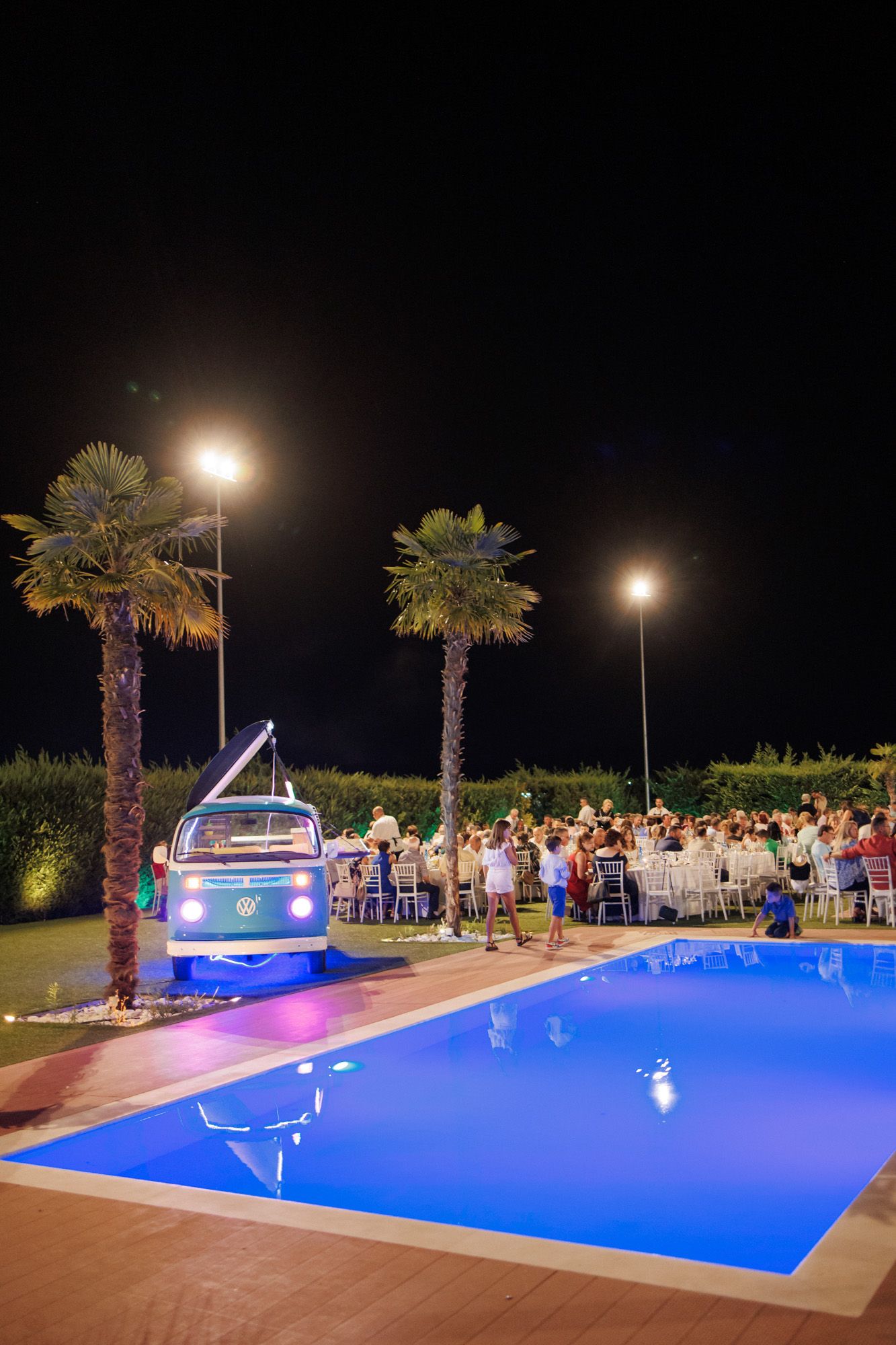 Nighttime outdoor wedding reception beside a blue pool with palm trees and a vintage VW bus DJ booth.