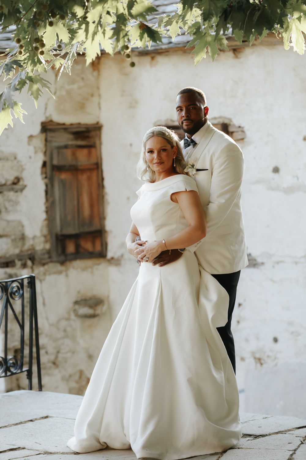 Interracial couple in wedding attire pose elegantly before a rustic stone wall with wooden shutters.