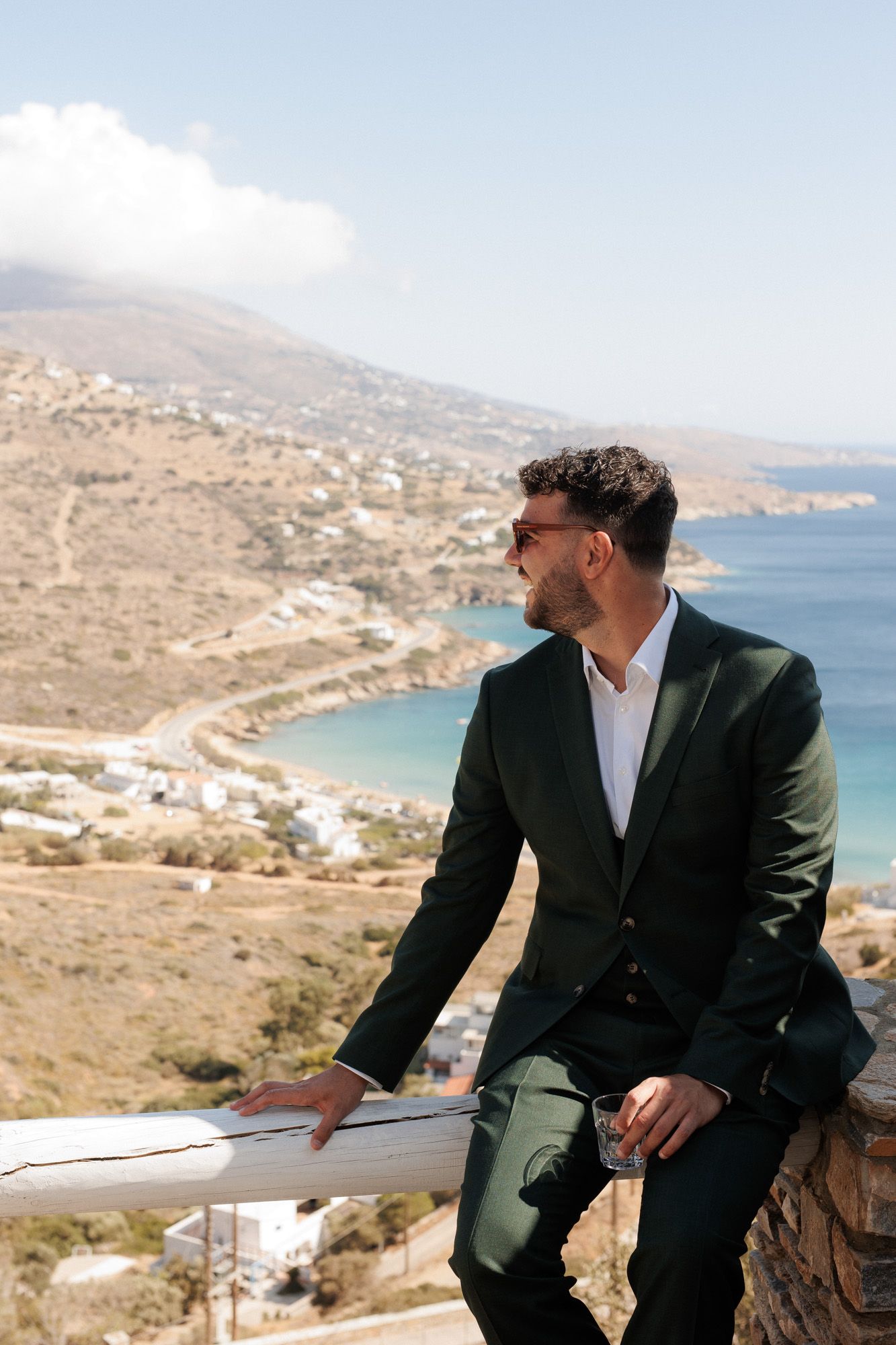 Elegant man in dark green suit leaning on railing overlooking scenic Greek island coastline.