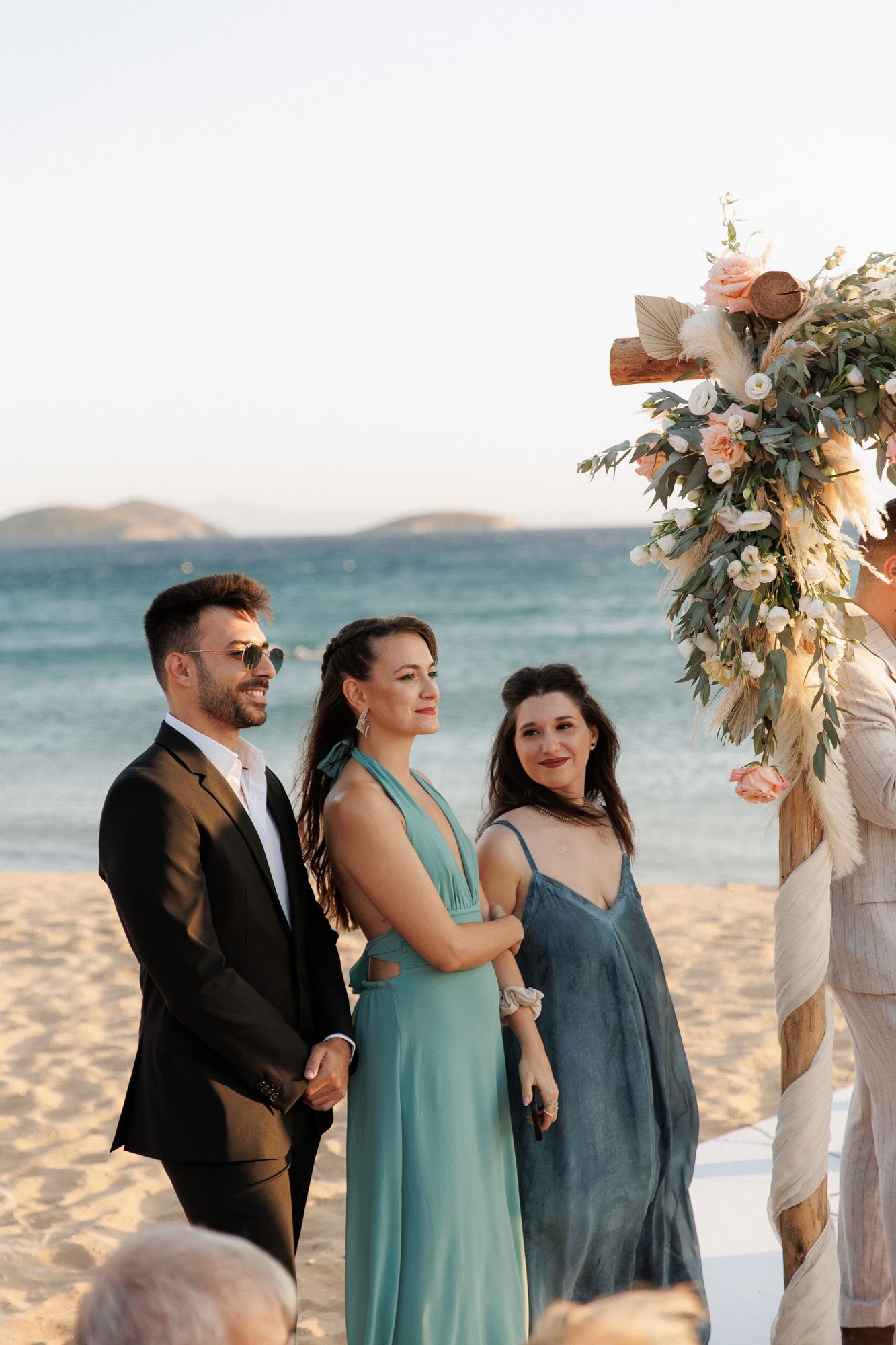 Wedding guests in elegant attire stand near floral arch at a sunny beachside ceremony.