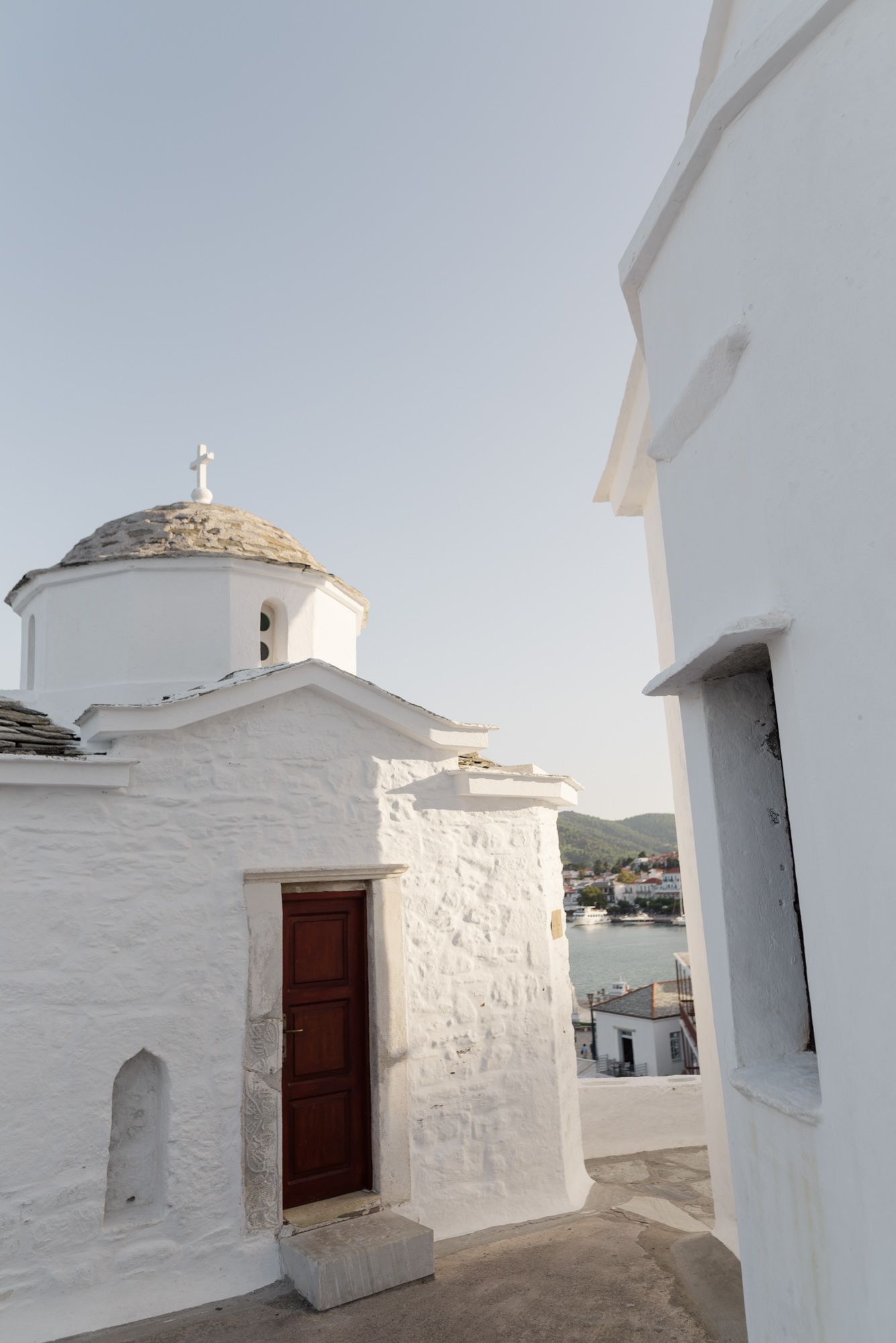 Traditional white-washed Greek chapel with domed roof and wooden door overlooking a coastal village.