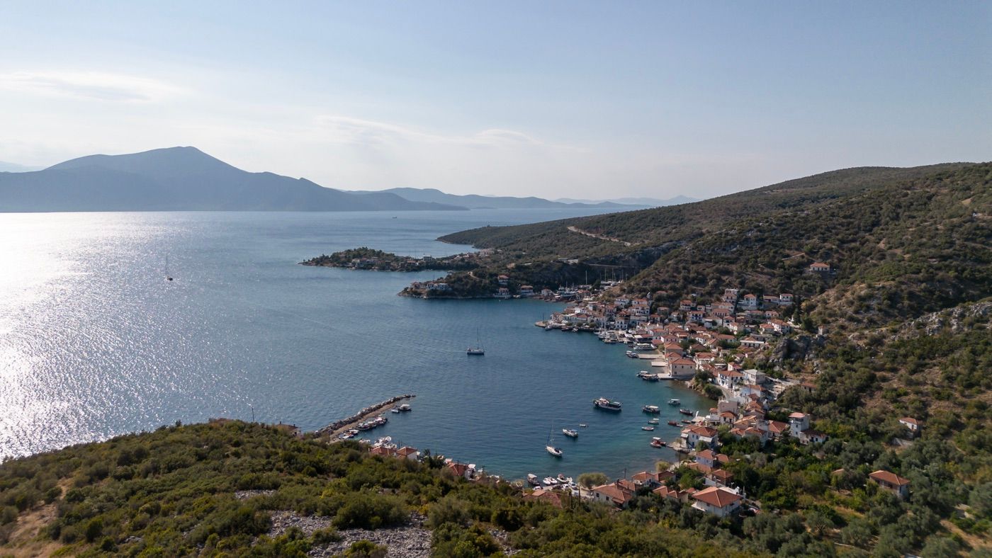 Aerial view of a picturesque harbor village nestled between green hills and sparkling blue water.