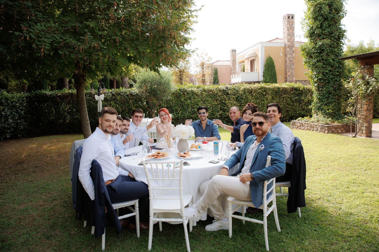 Group of well-dressed groomsmen and guests seated at an outdoor white table in a lush garden.