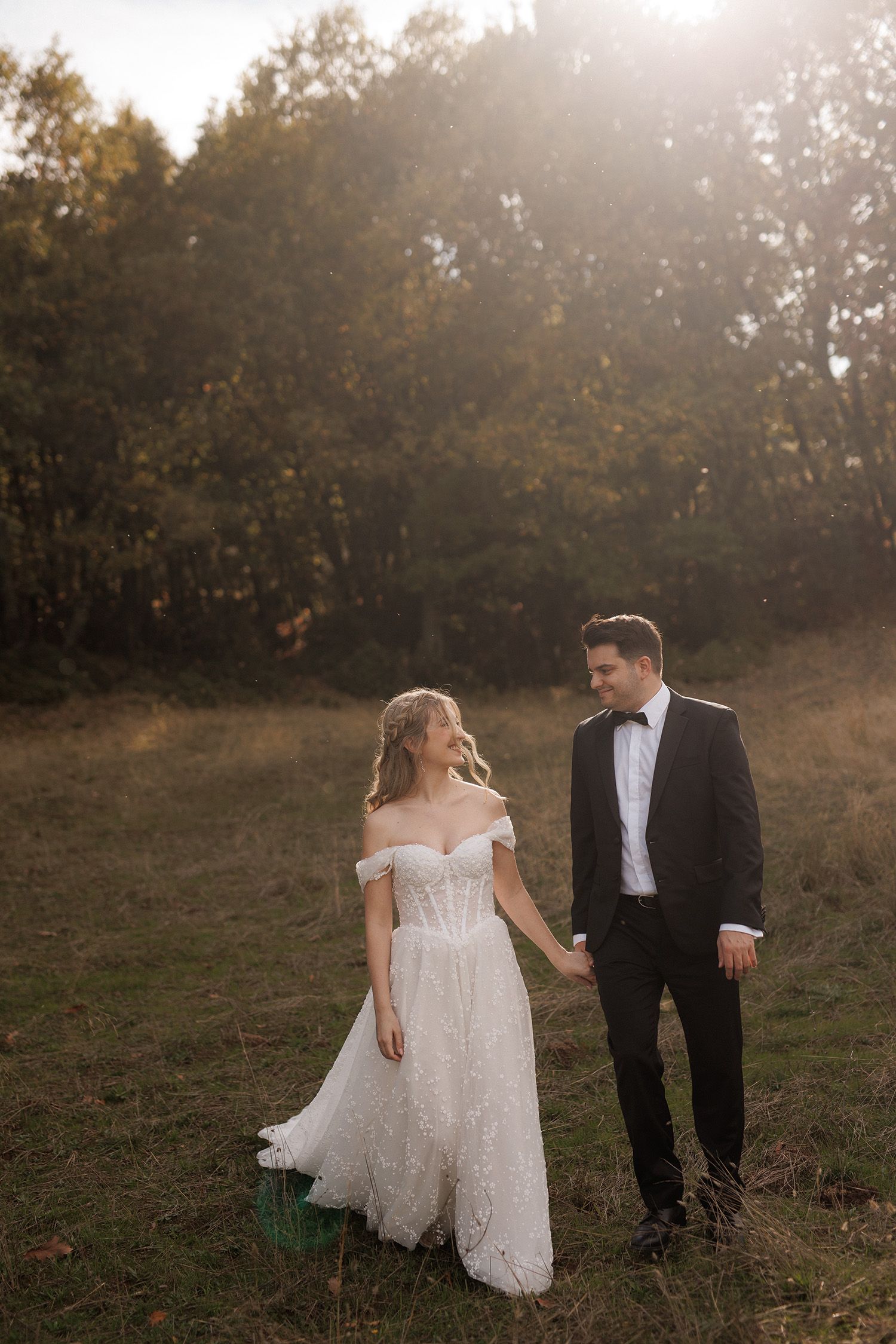 A bride in a white off-shoulder gown and groom in black tuxedo hold hands in a sunlit autumn meadow.
