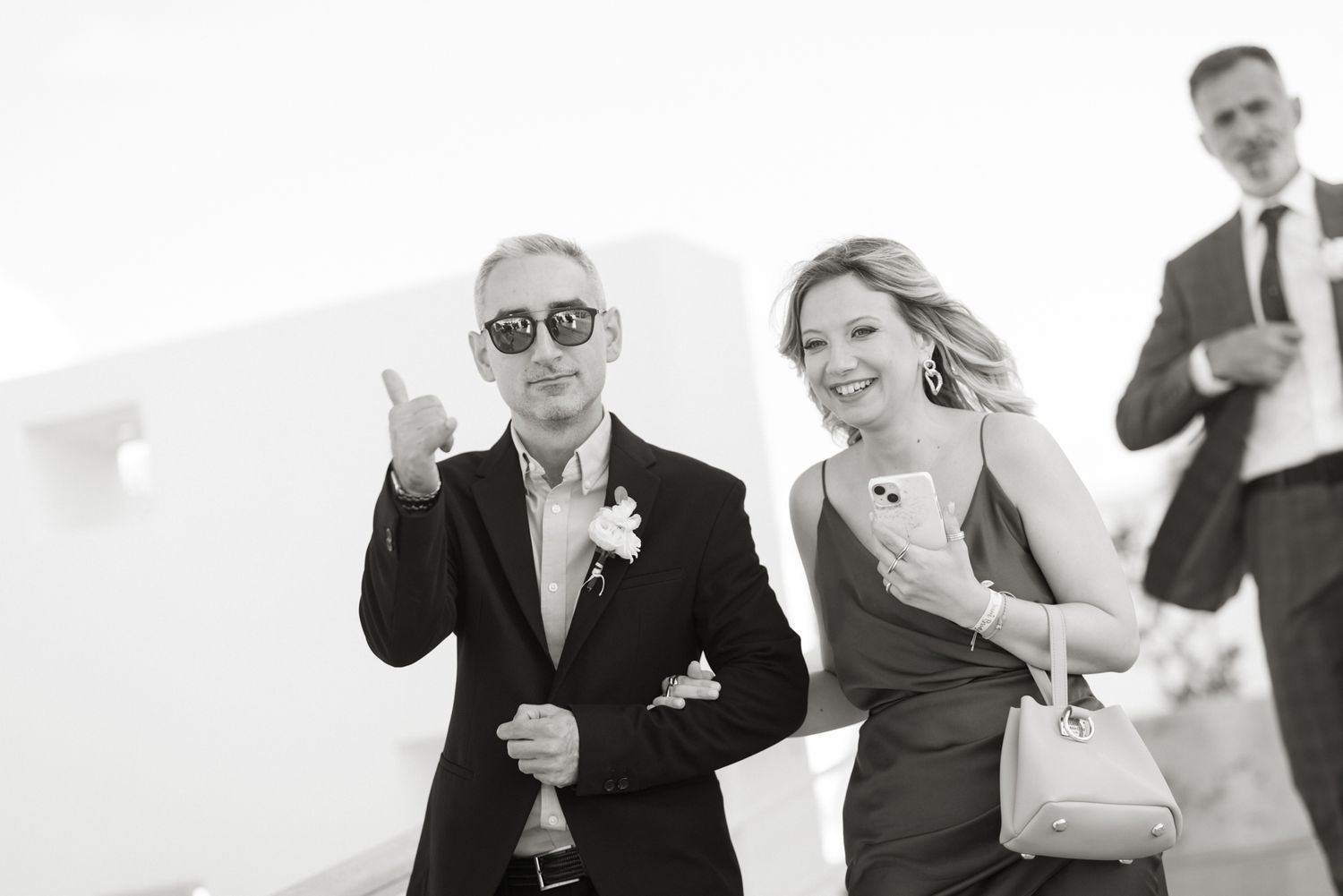 Black and white photo of smiling wedding guests arriving, man giving thumbs up at outdoor venue.