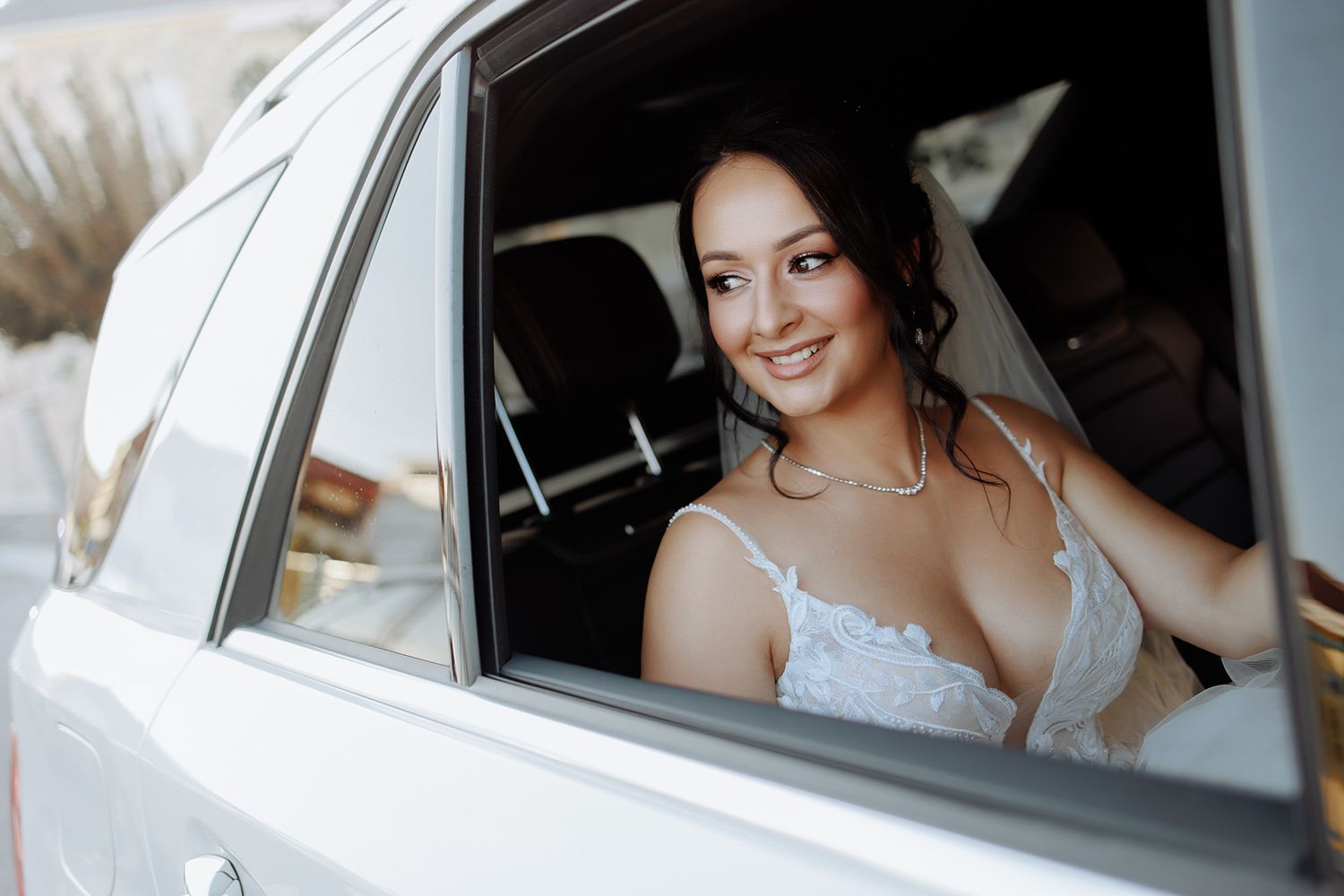 A glowing bride smiles from inside a white wedding car wearing a delicate lace gown and sparkling necklace.