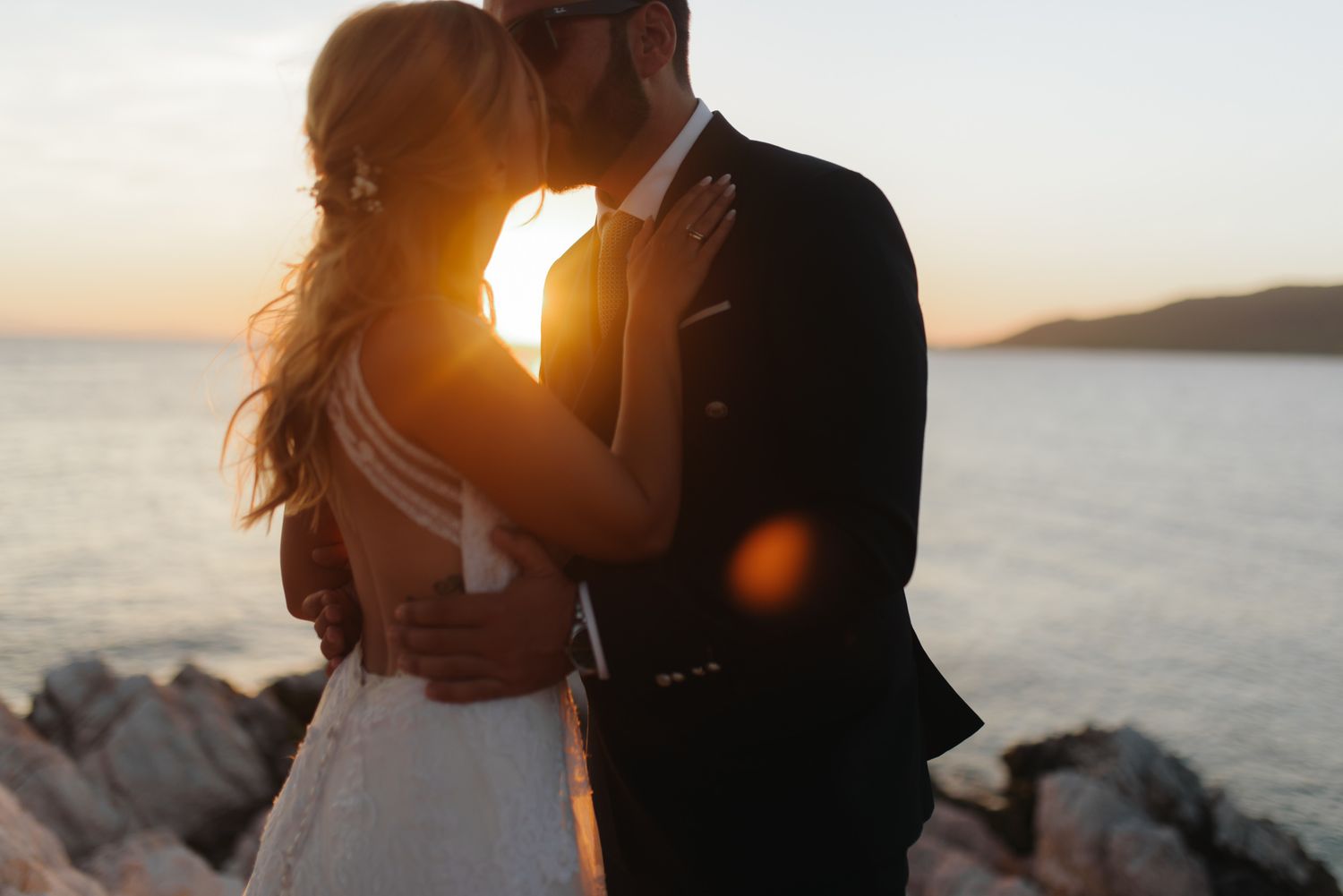 Bride and groom sharing romantic sunset kiss on rocky seaside with golden backlight.