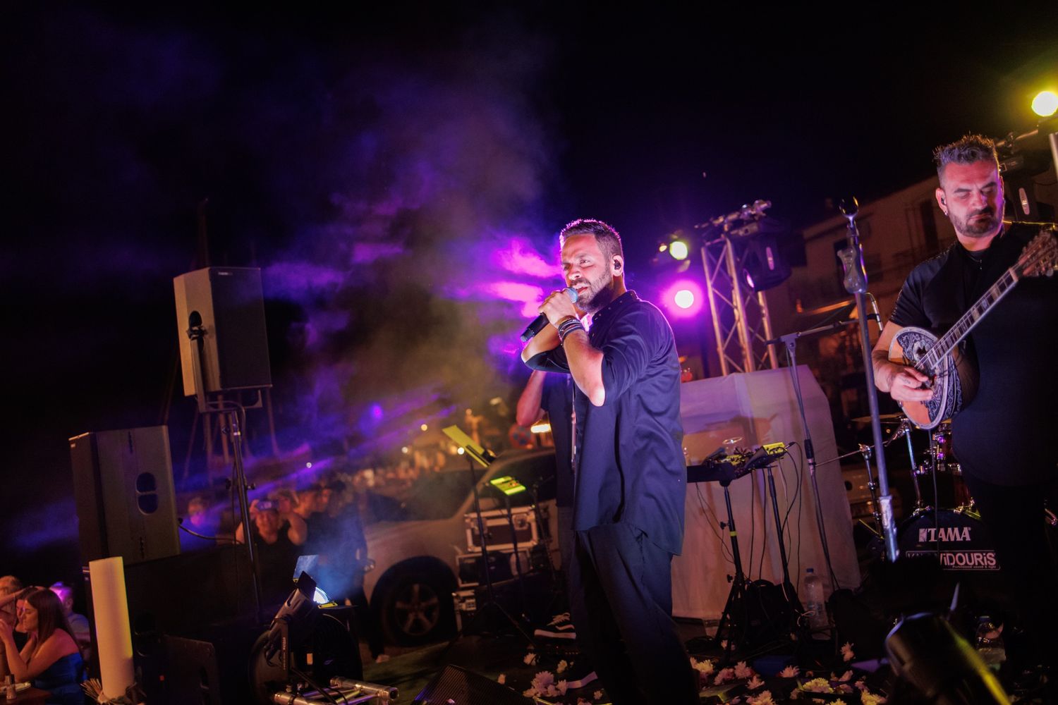 Live band performing on a lit stage at night with purple smoke and crowd below.