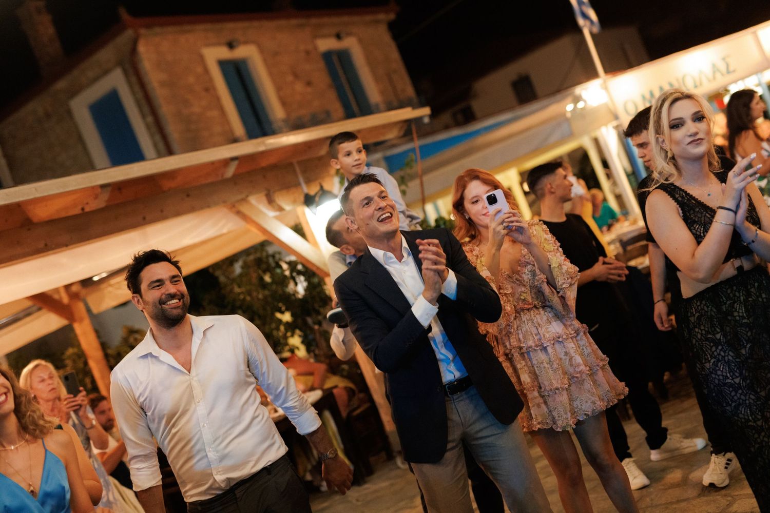 Wedding guests laughing and celebrating at a festive outdoor evening reception with warm lights.